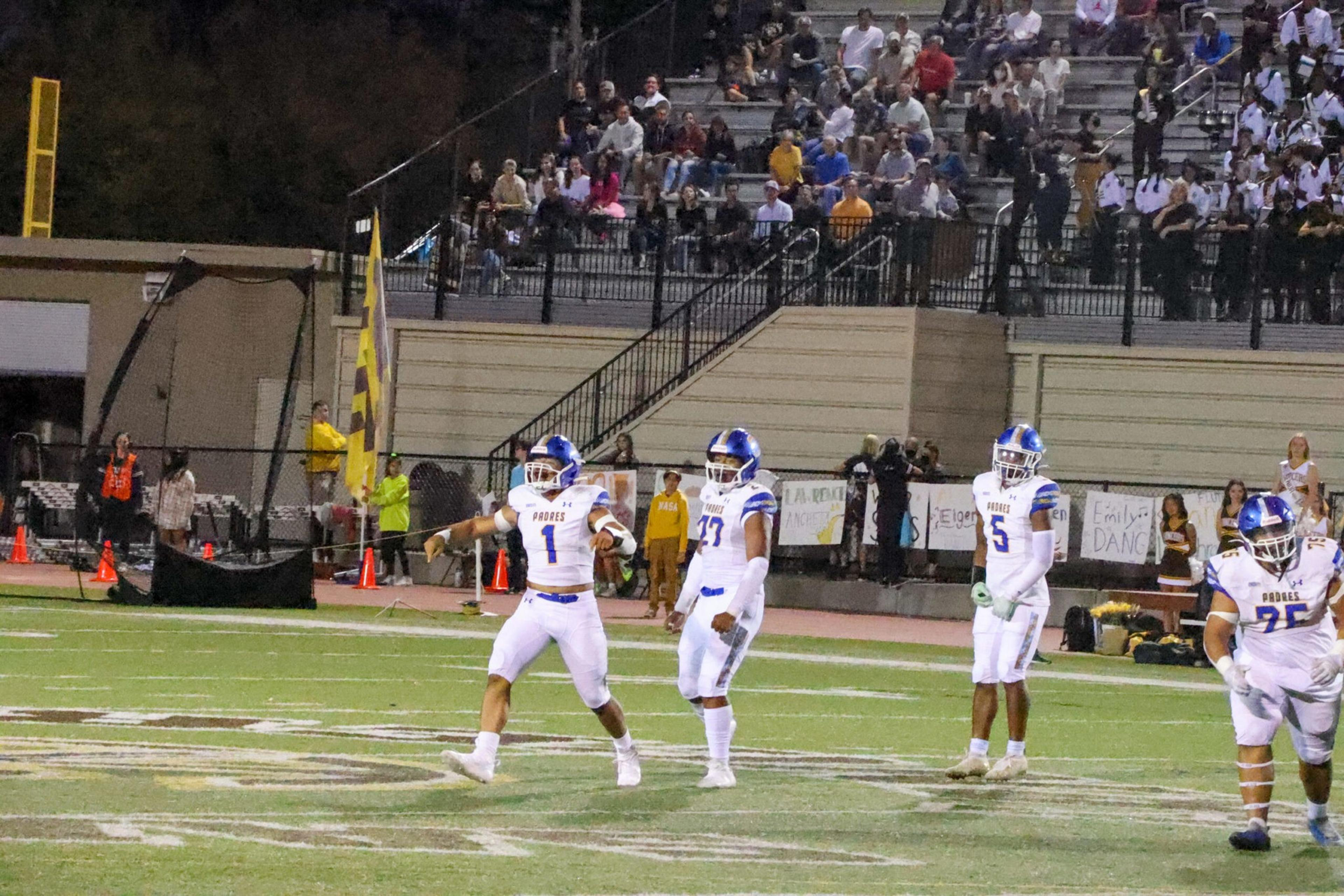 Four football players in white and blue uniforms stand on a field, with a crowd of spectators seated on bleachers behind them at night.