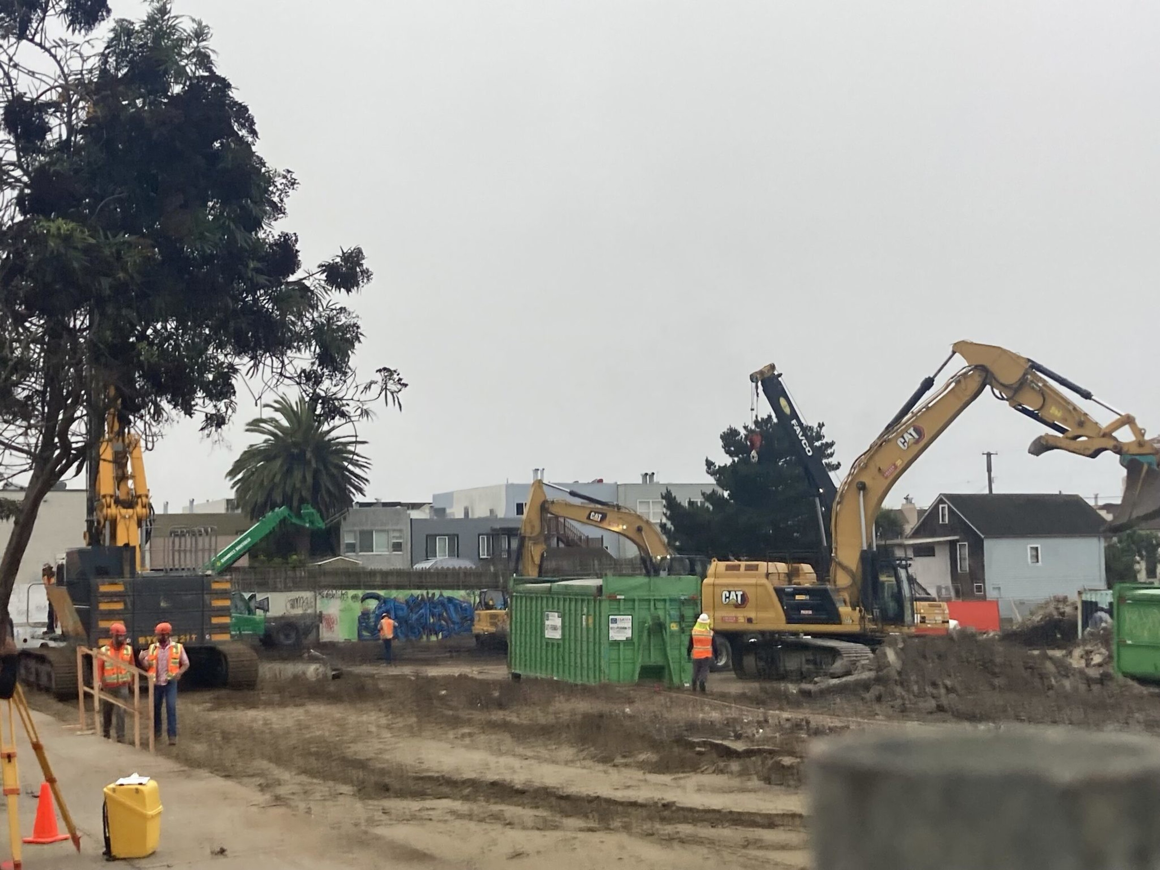 Construction workers in orange vests and helmets operate heavy machinery, including excavators, amid dirt and green dumpsters at a building site near houses.