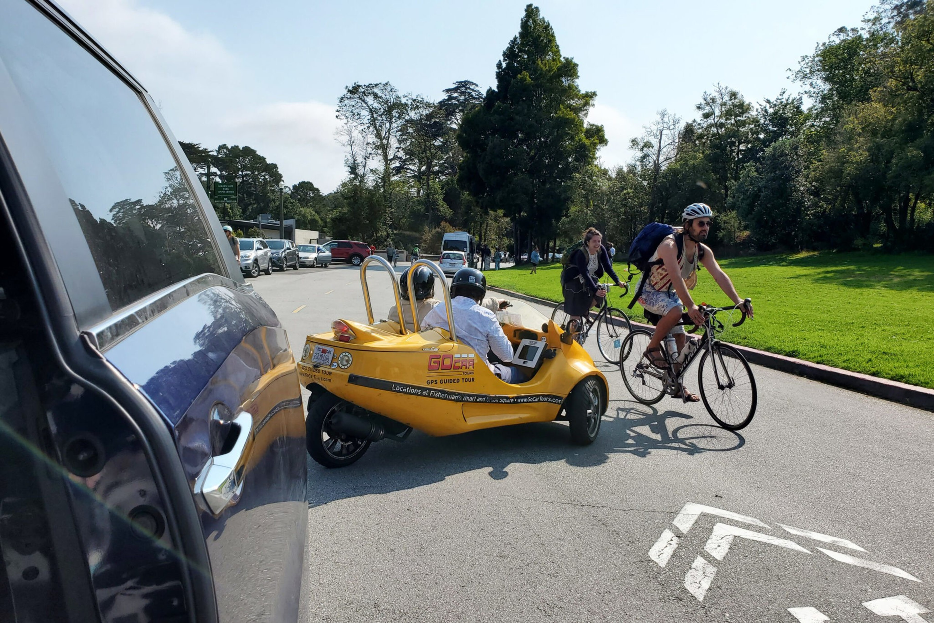A yellow three-wheeled go-kart with two people rides past two cyclists on a sunny road with trees and grass nearby, with cars lined up in the background.
