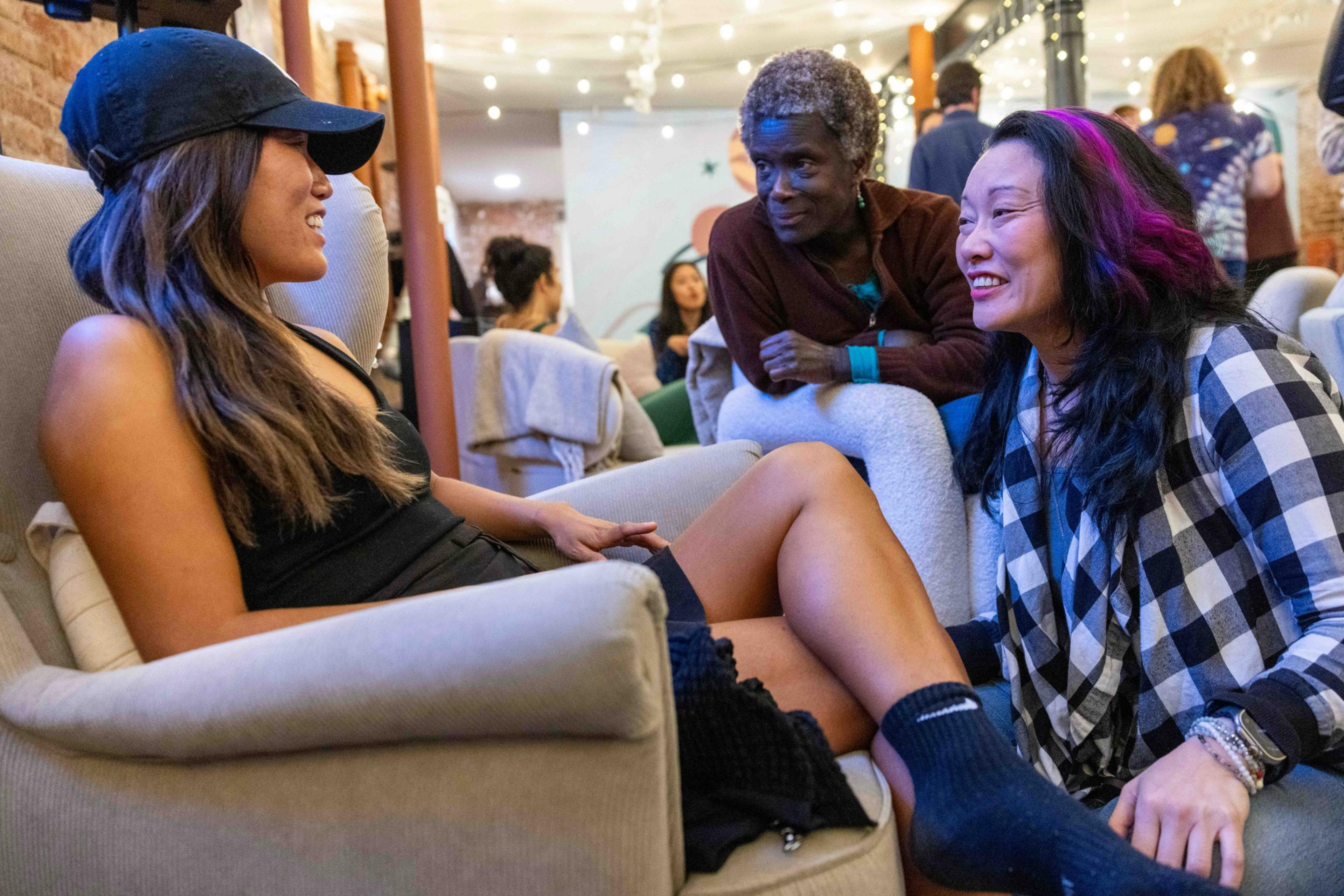 Three people are engaged in a lively conversation in a cozy, well-lit room; one woman sits in a chair while the other two kneel nearby.