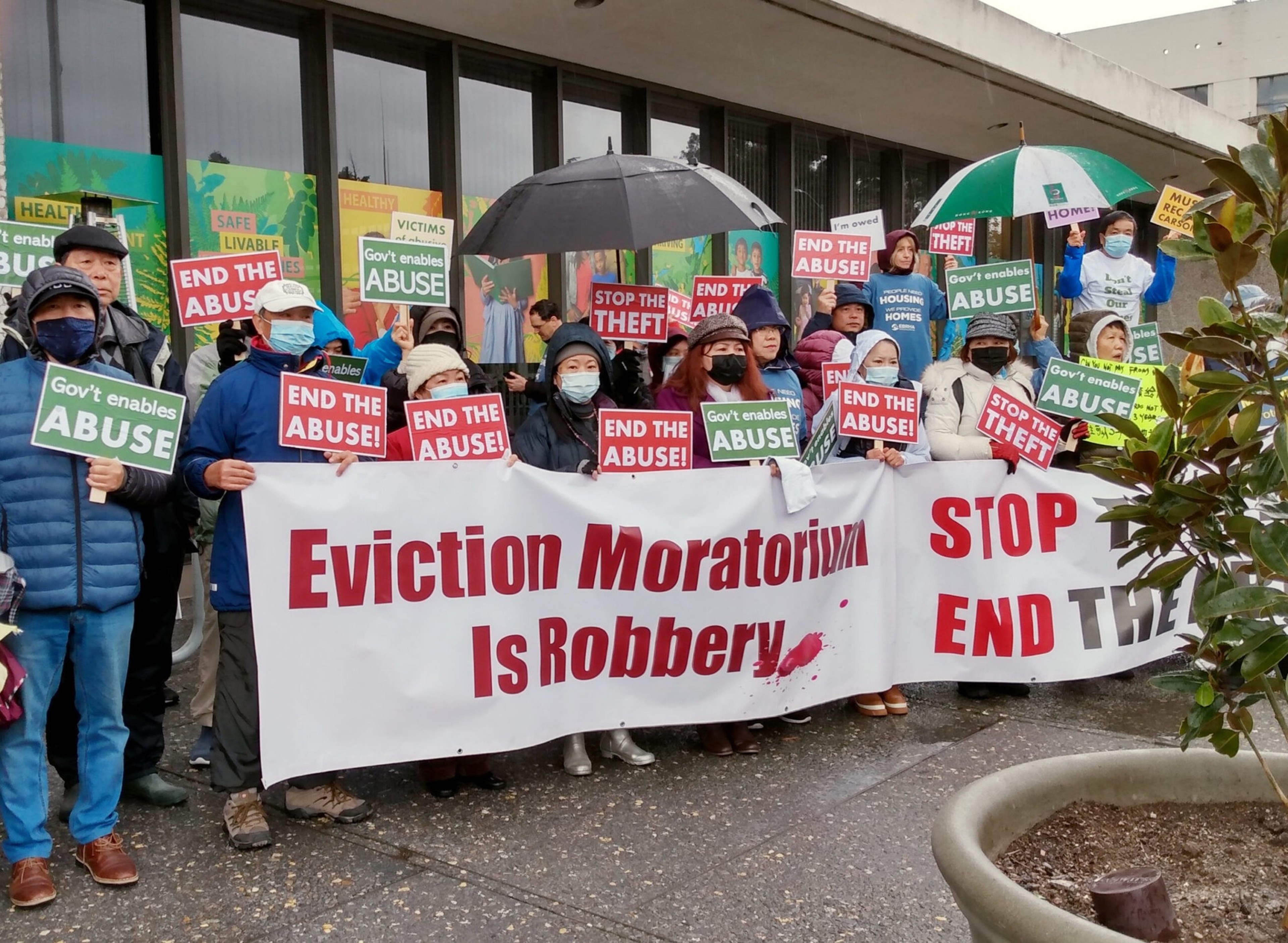 A diverse group of protesters holding signs that say "END THE ABUSE!", "Gov't enables ABUSE," and a large banner reading "Eviction Moratorium Is Robbery."