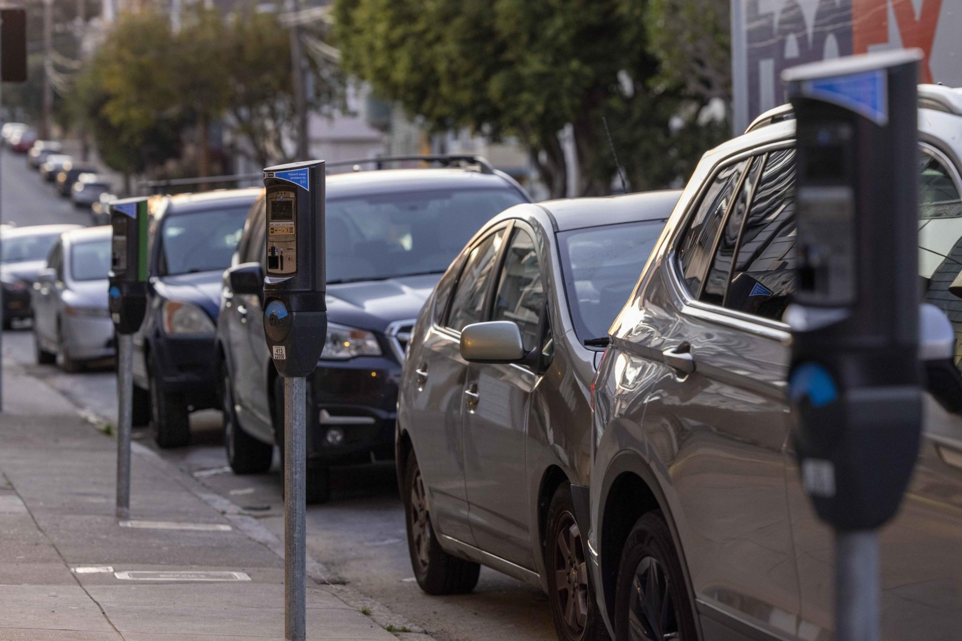 Cars are parked along a street with several parking meters visible. Trees and more cars are in the background, creating a typical urban scene.