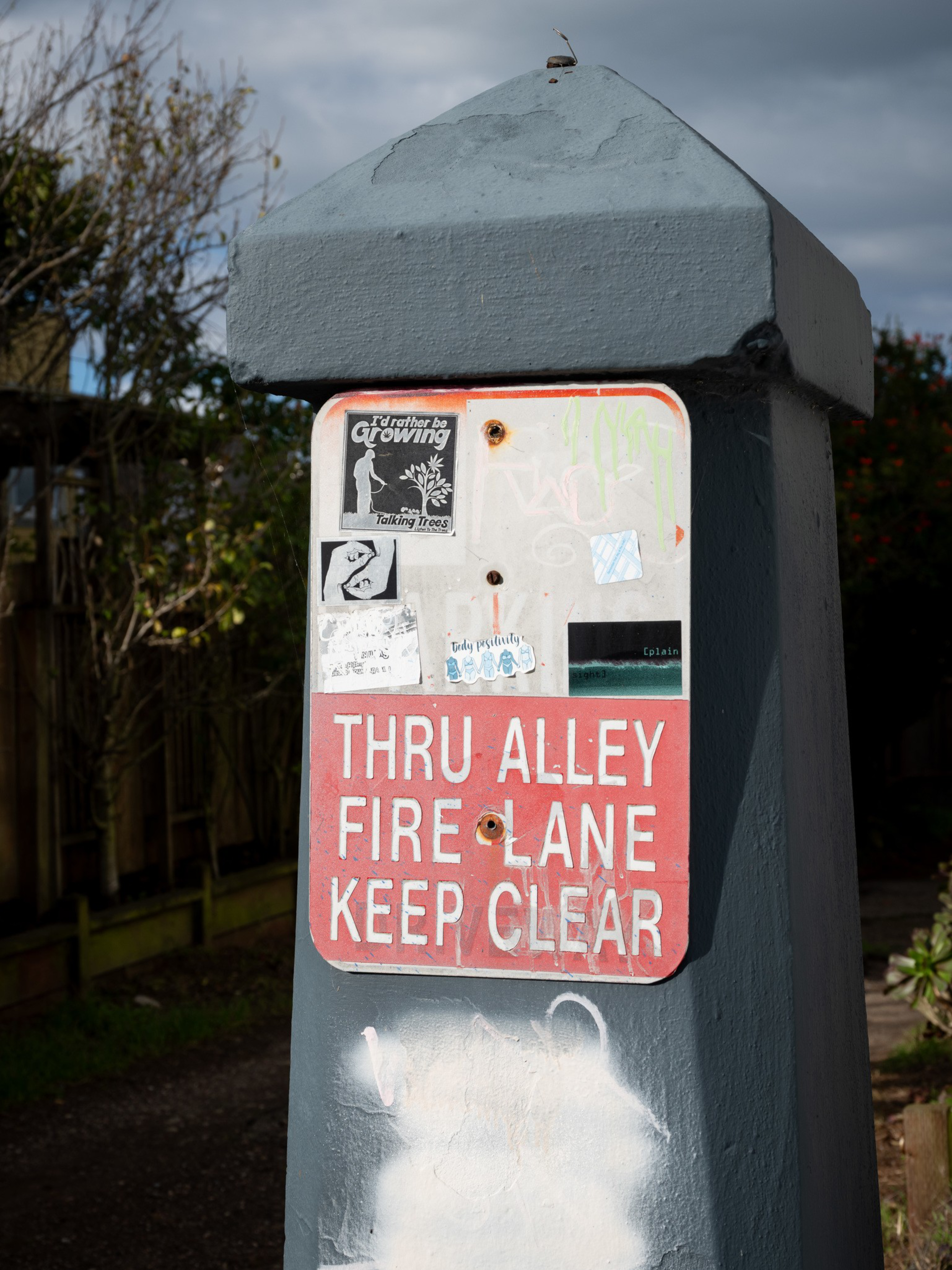A gray post displays a worn white and red sign reading “THRU ALLEY FIRE LANE KEEP CLEAR,” covered with various small stickers.