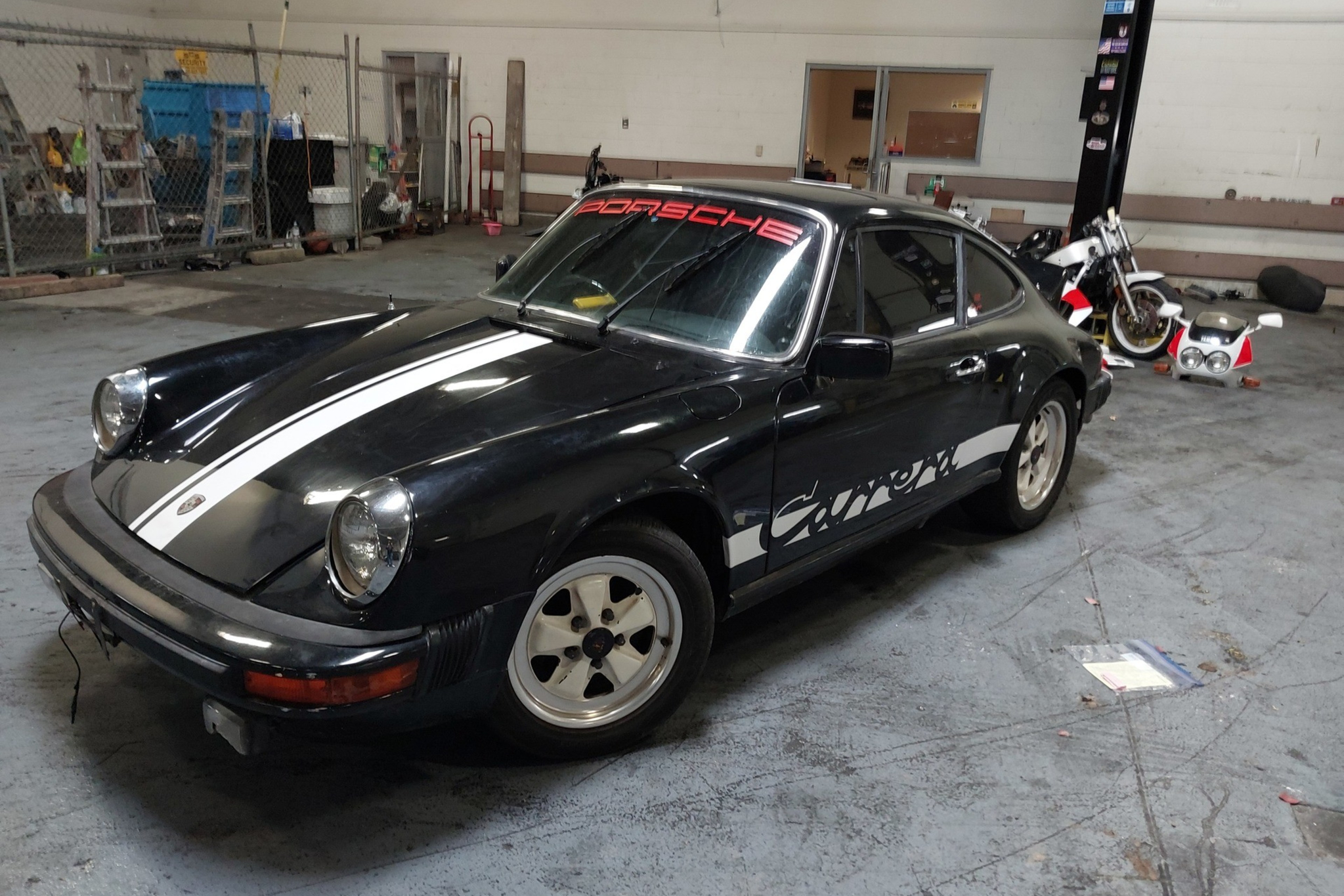 A black Porsche Carrera with white racing stripes and decals is parked inside a garage, surrounded by tools and a motorcycle.