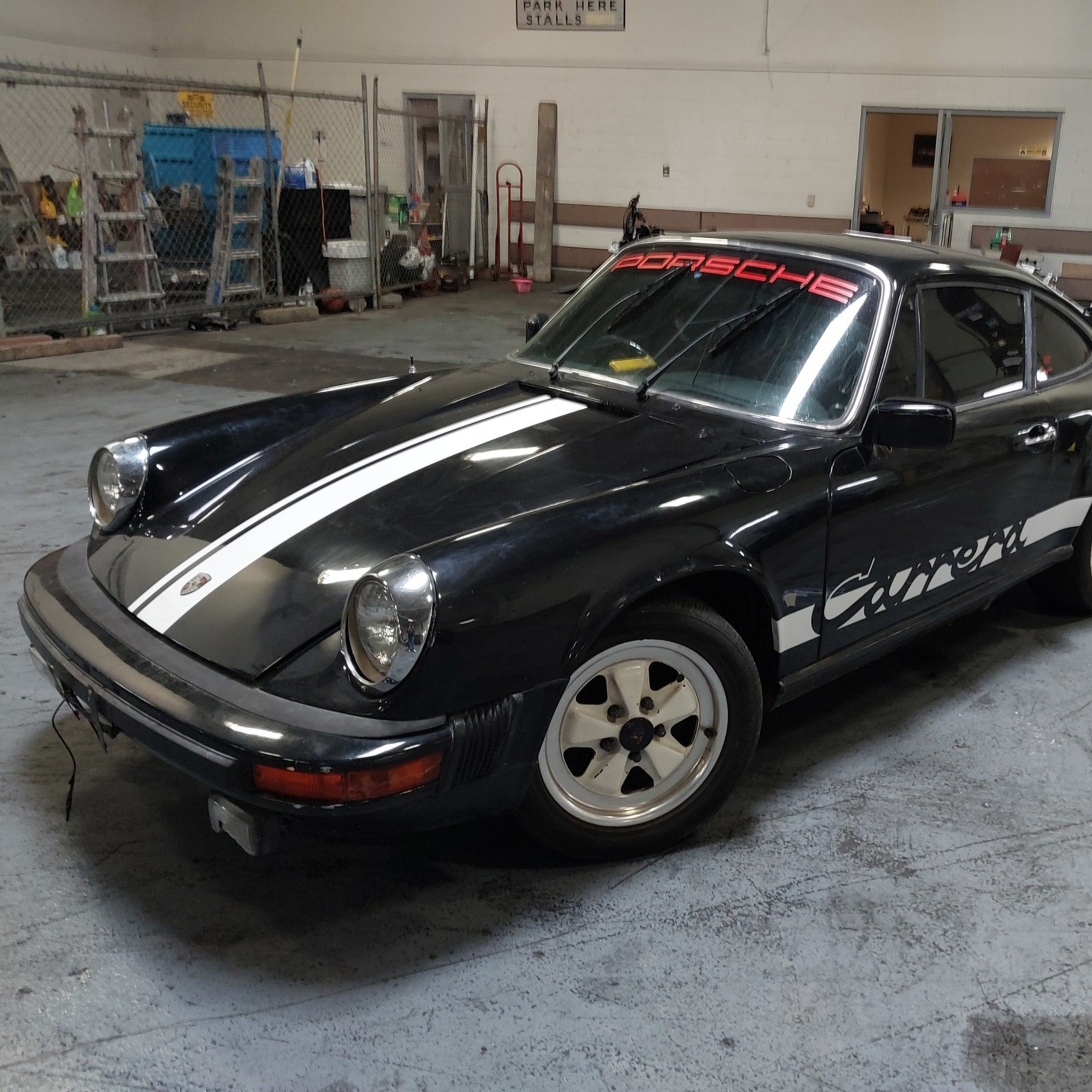 A black Porsche Carrera with white racing stripes and decals is parked inside a garage, surrounded by tools and a motorcycle.