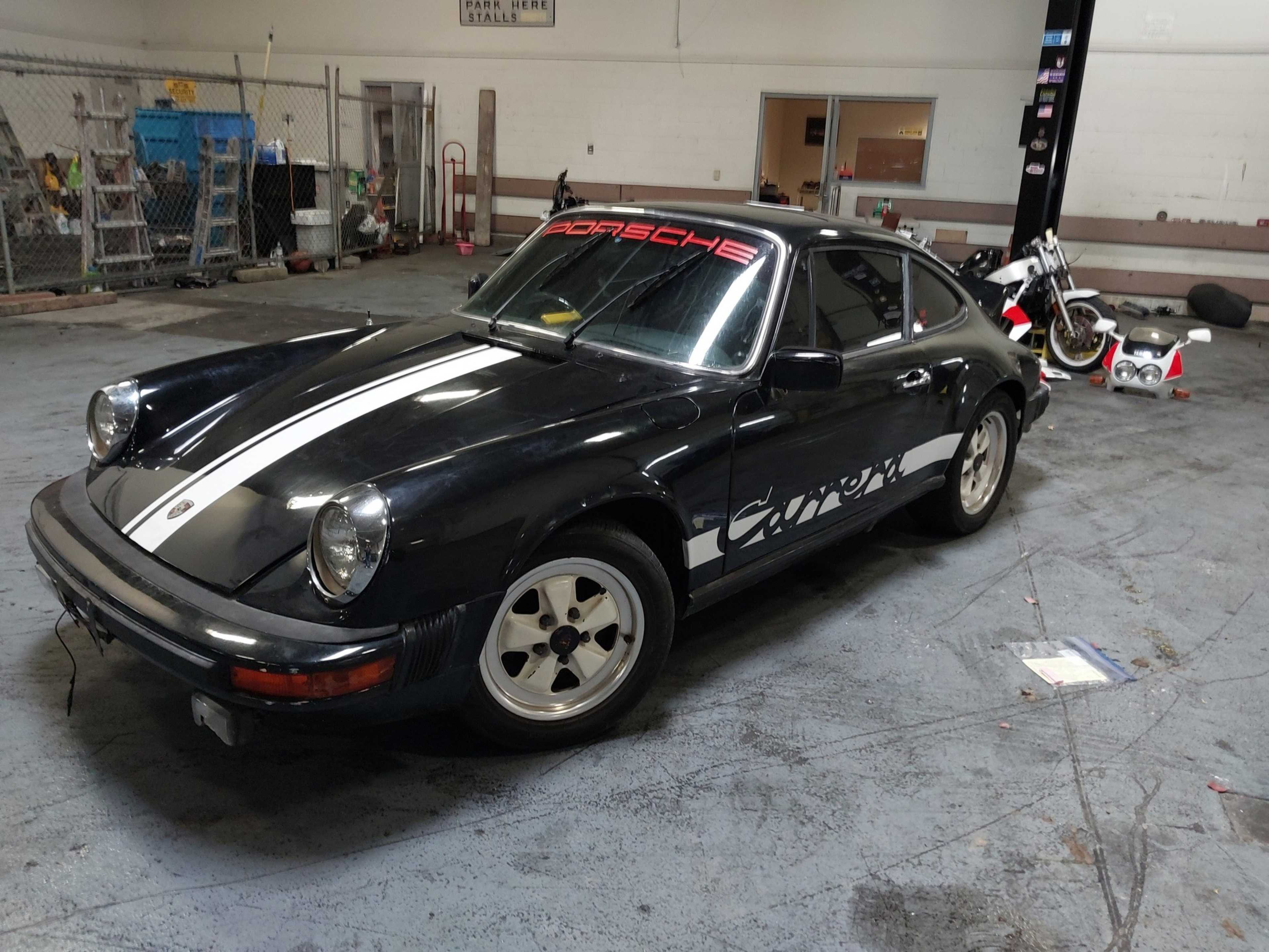 A black Porsche Carrera with white racing stripes and decals is parked inside a garage, surrounded by tools and a motorcycle.