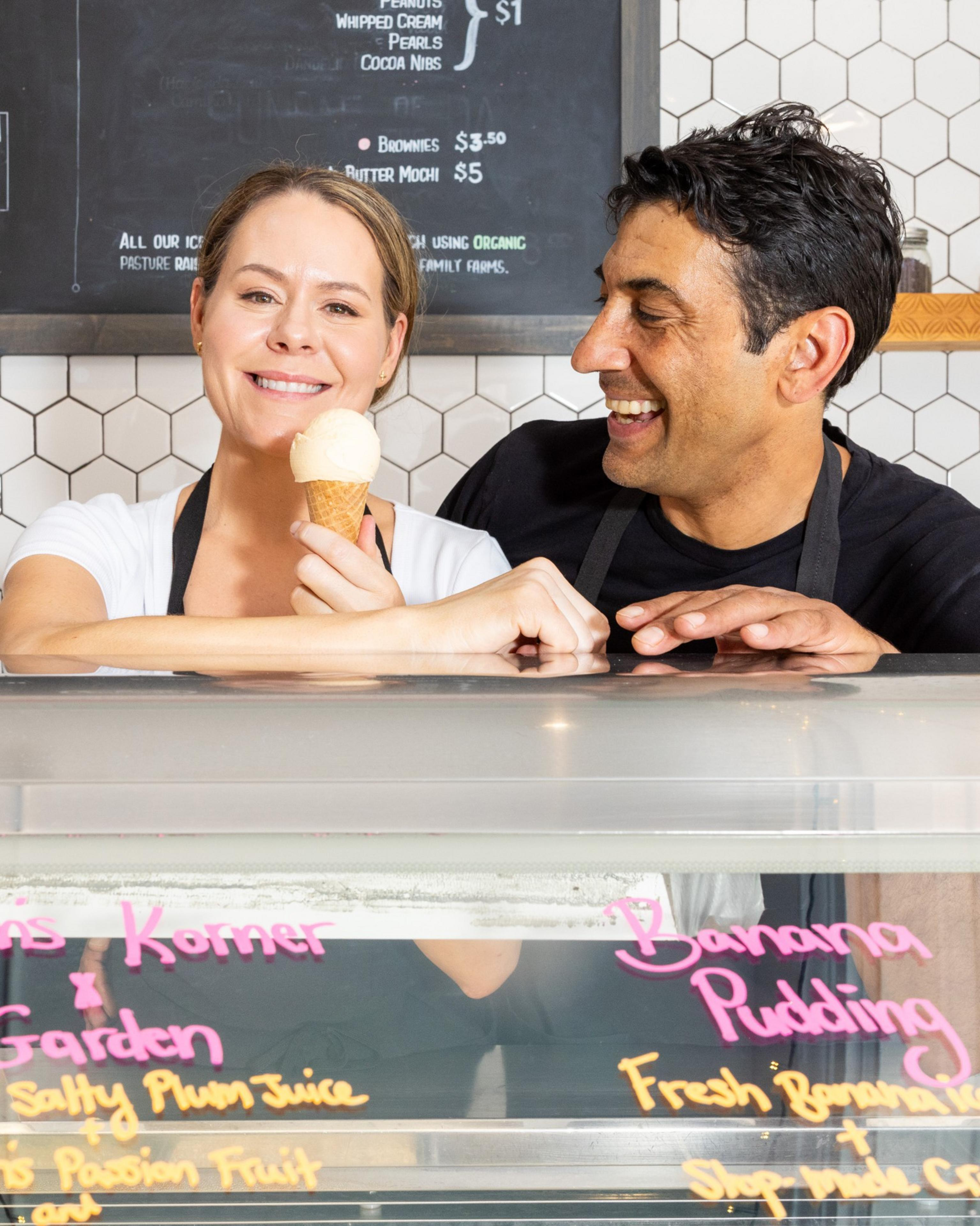 Two people behind an ice cream counter smile, with one holding an ice cream cone, in front of a menu on a hexagonal tile background.