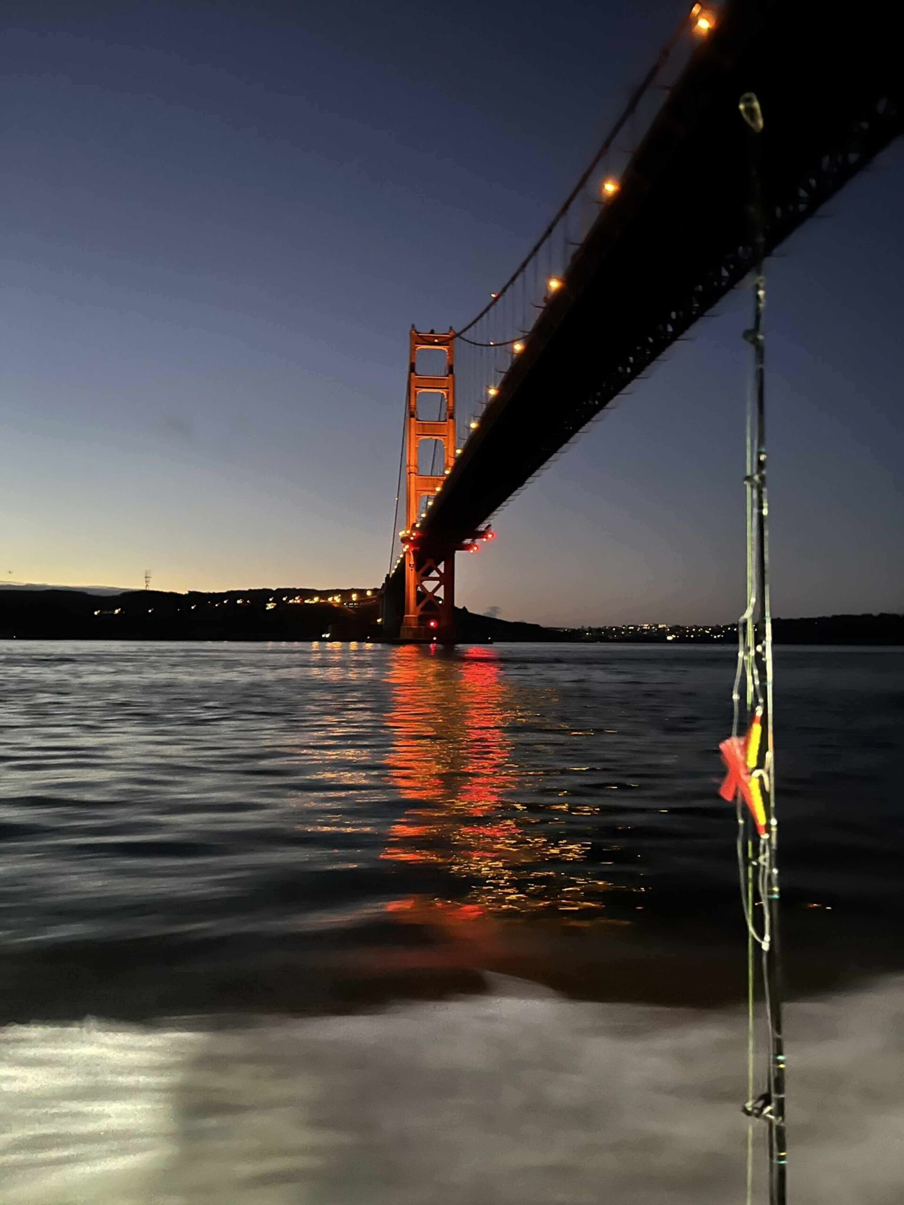 The illuminated Golden Gate Bridge stretches across calm waters at dusk, reflecting orange lights, with part of a fishing rod visible in the foreground.