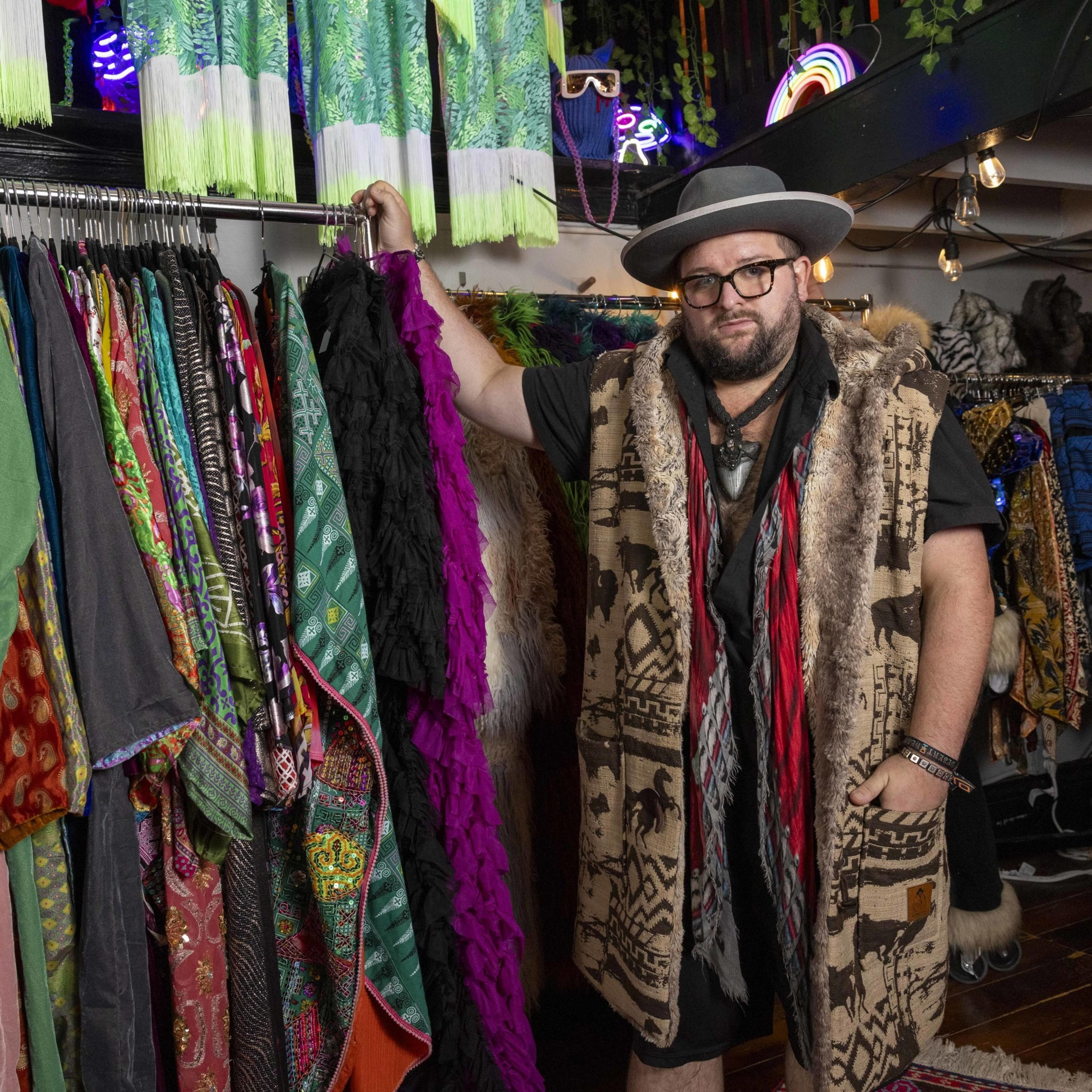 A man wearing a wide-brimmed hat and a patterned fur-trimmed vest stands in a colorful clothing store, holding a black ruffled garment and surrounded by vibrant fabrics.