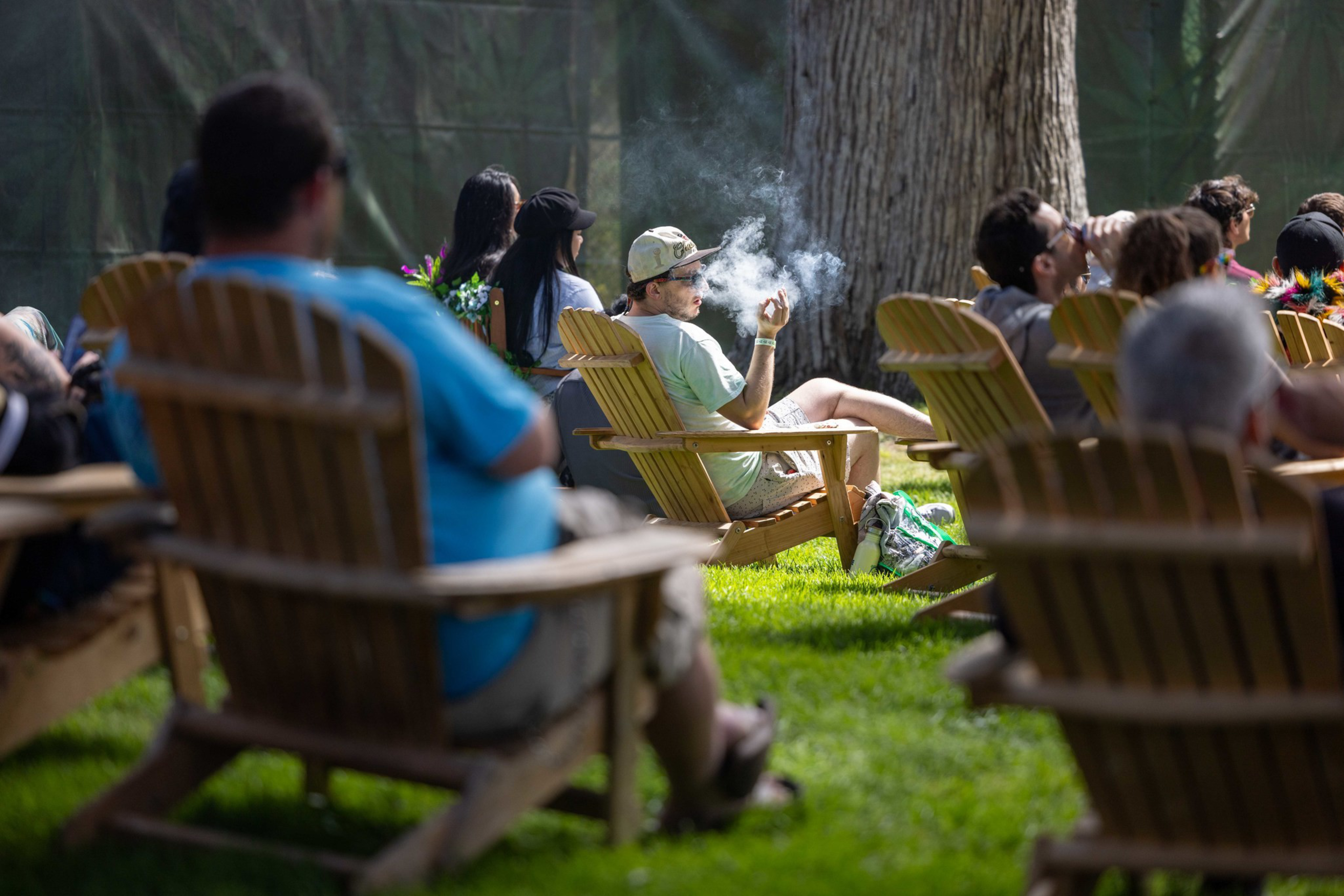 People sit in wooden Adirondack chairs on grass, with one person in the center smoking and exhaling smoke in an outdoor setting.