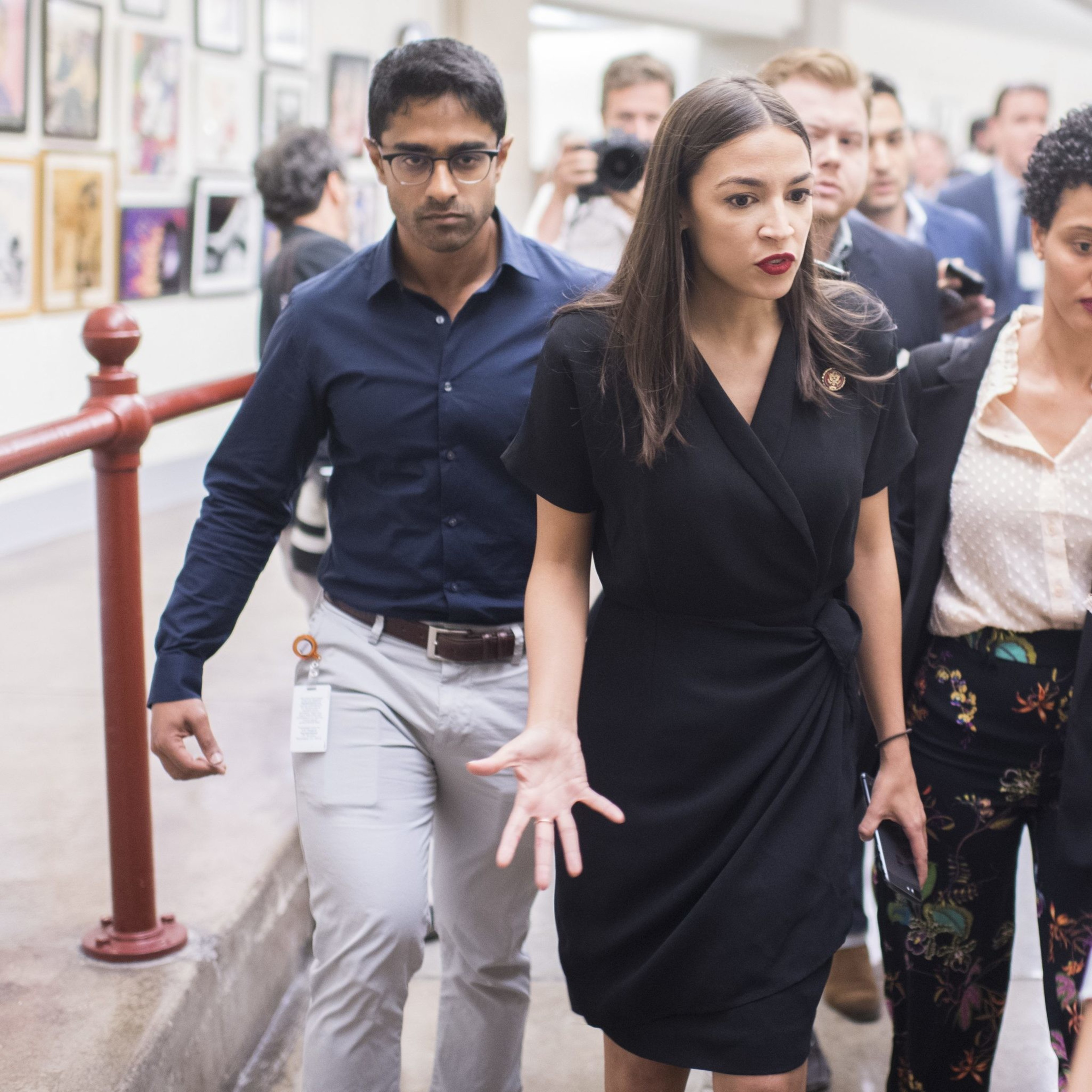 A woman in a black dress walks down a hallway, looking focused, surrounded by people. One man holds a camera, and framed pictures hang on the wall.