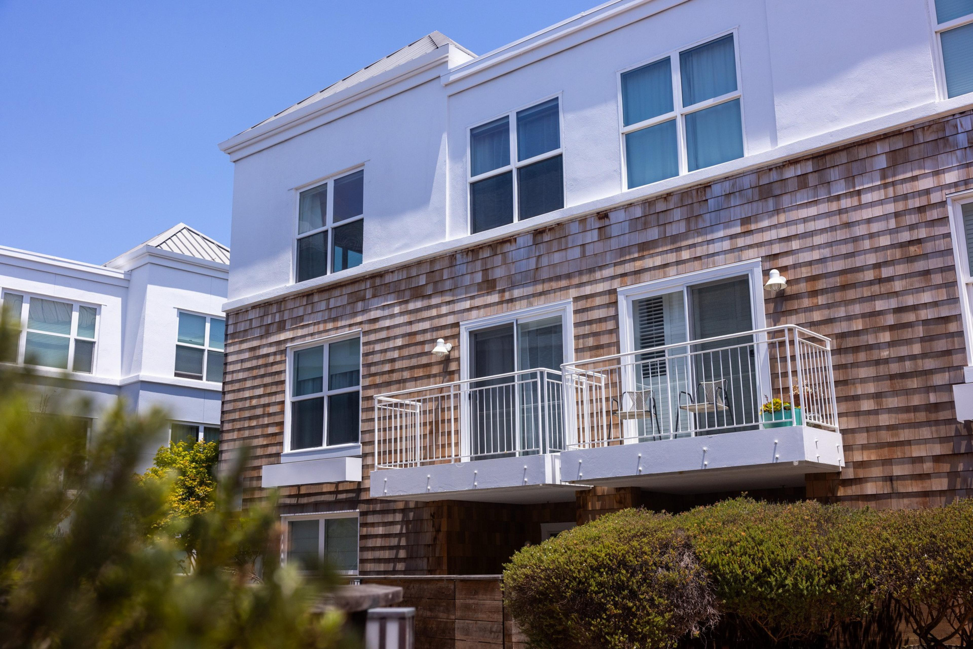 The image shows a modern apartment building with wooden siding and white trim. There are balconies with white railings, large windows, and plants in front.