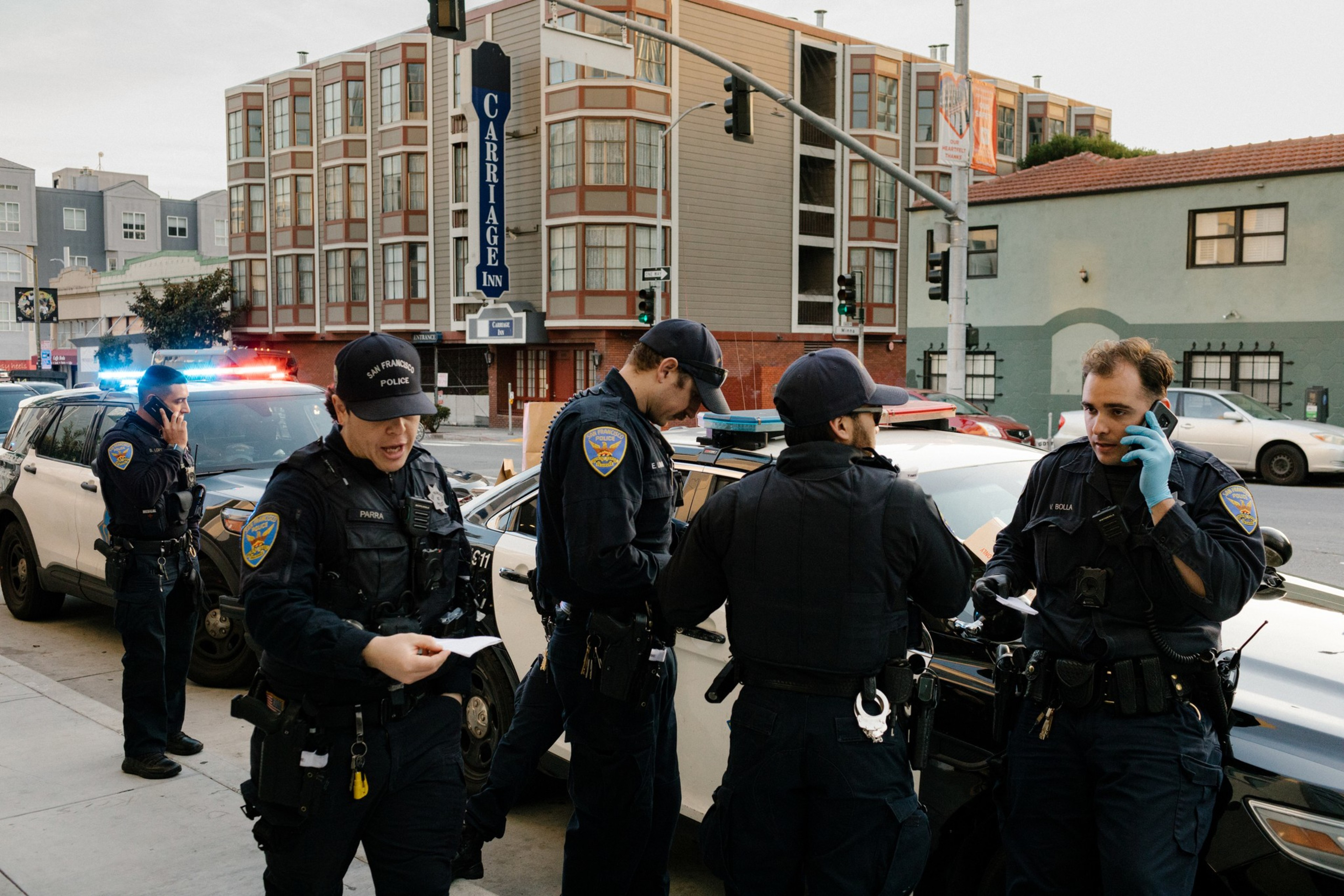 Four police officers are gathered near patrol cars with flashing lights. The officers are engaged in discussions, phone calls, and examining documents. In the background, there is a "Carriage Inn" building.