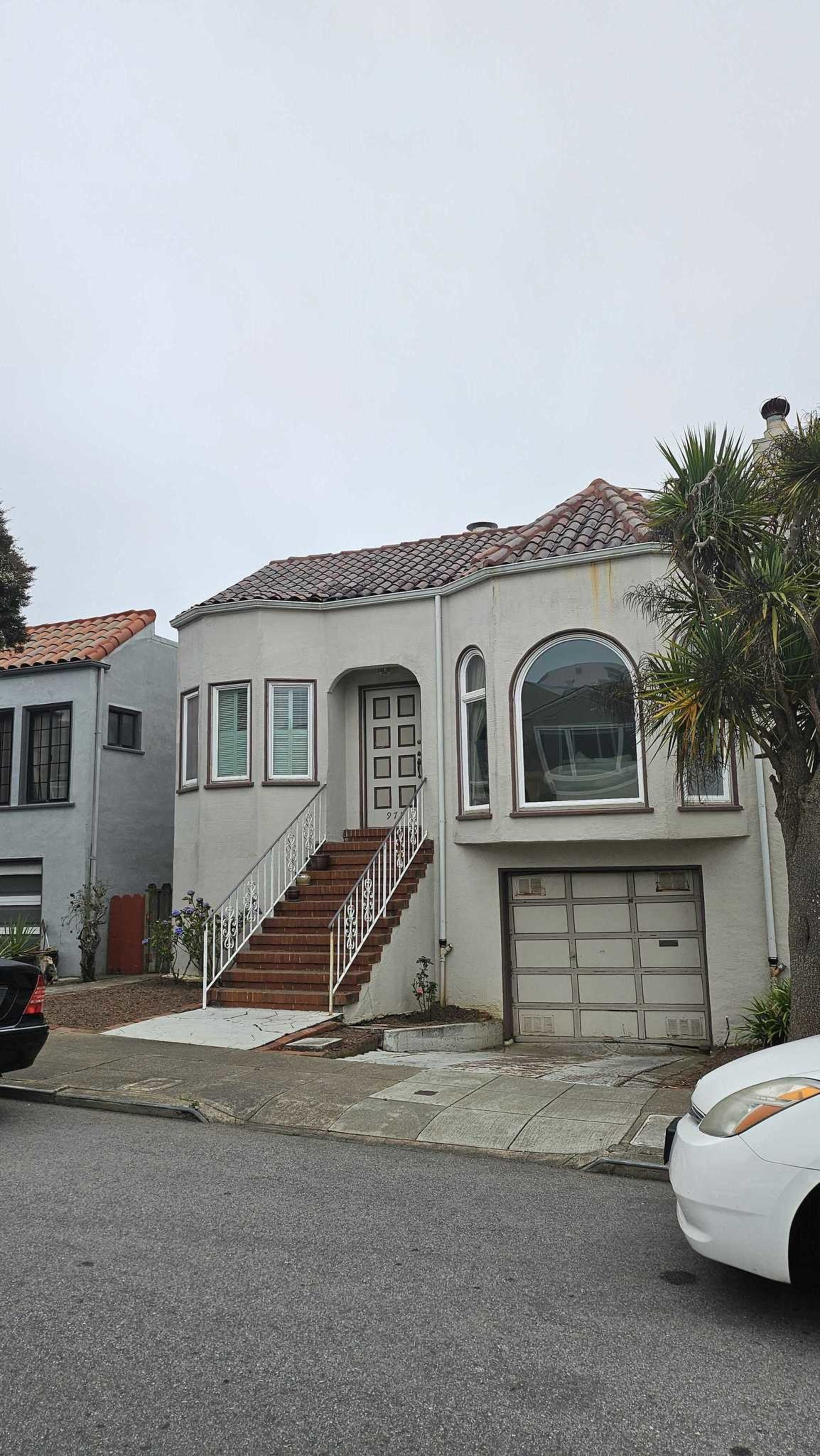 A two-story beige house with a tile roof features a staircase leading to the front door, a garage below, large arched windows, and a palm tree nearby.