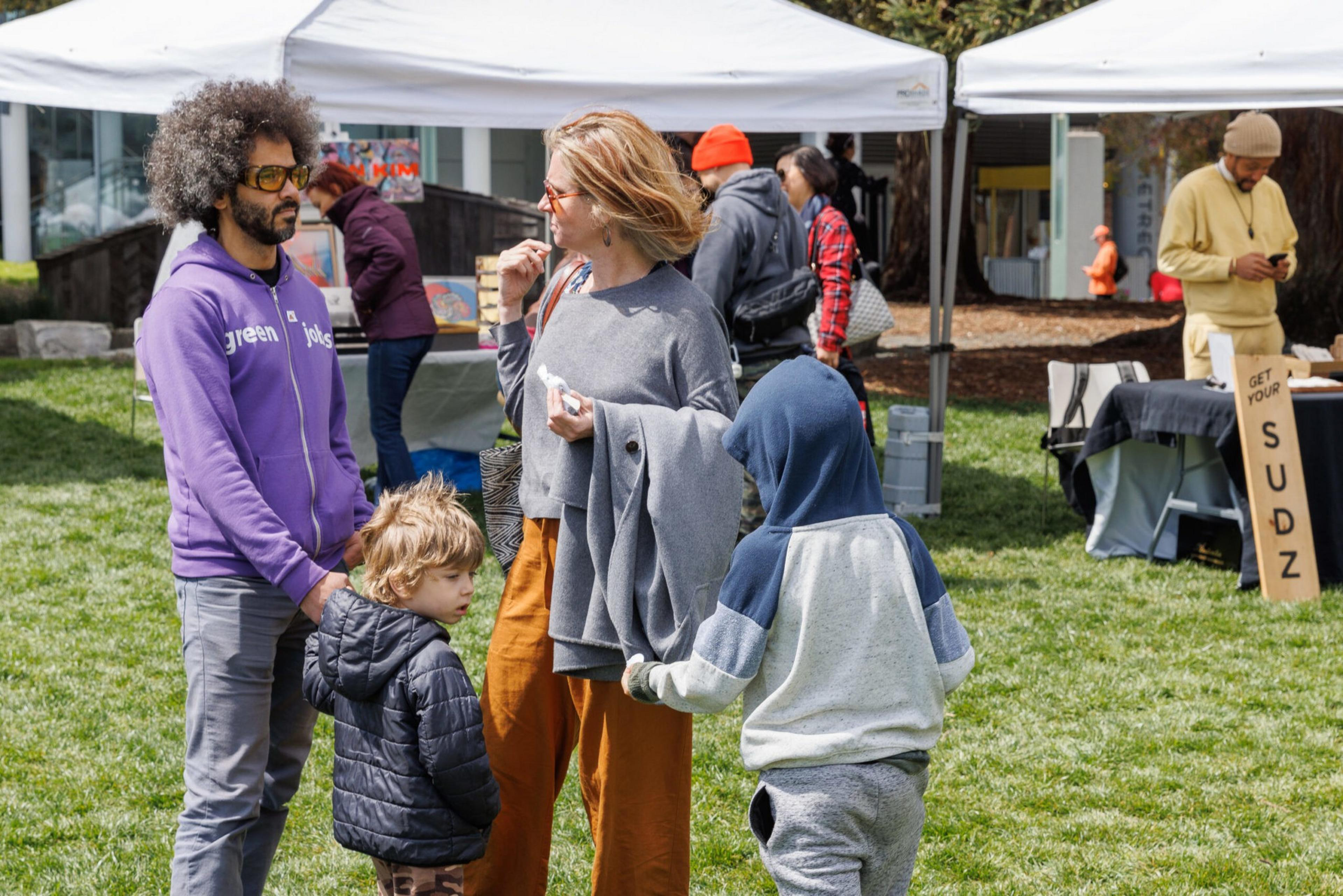 A family is conversing at an outdoor market with tents and other visitors in the background.