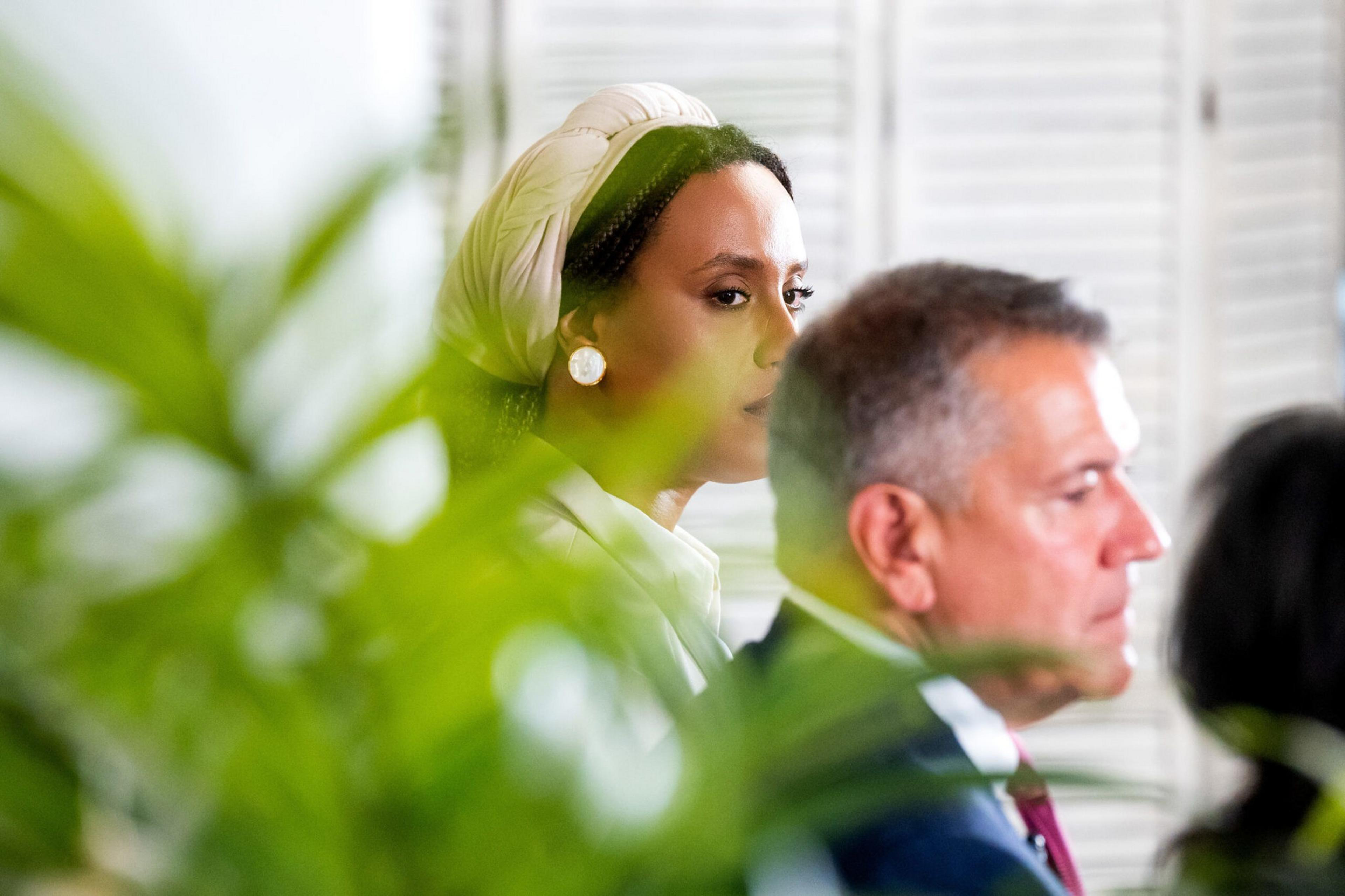 A woman wearing a white headscarf and pearl earrings looks ahead, partially obscured by green foliage, while a man in a suit sits in front of her.