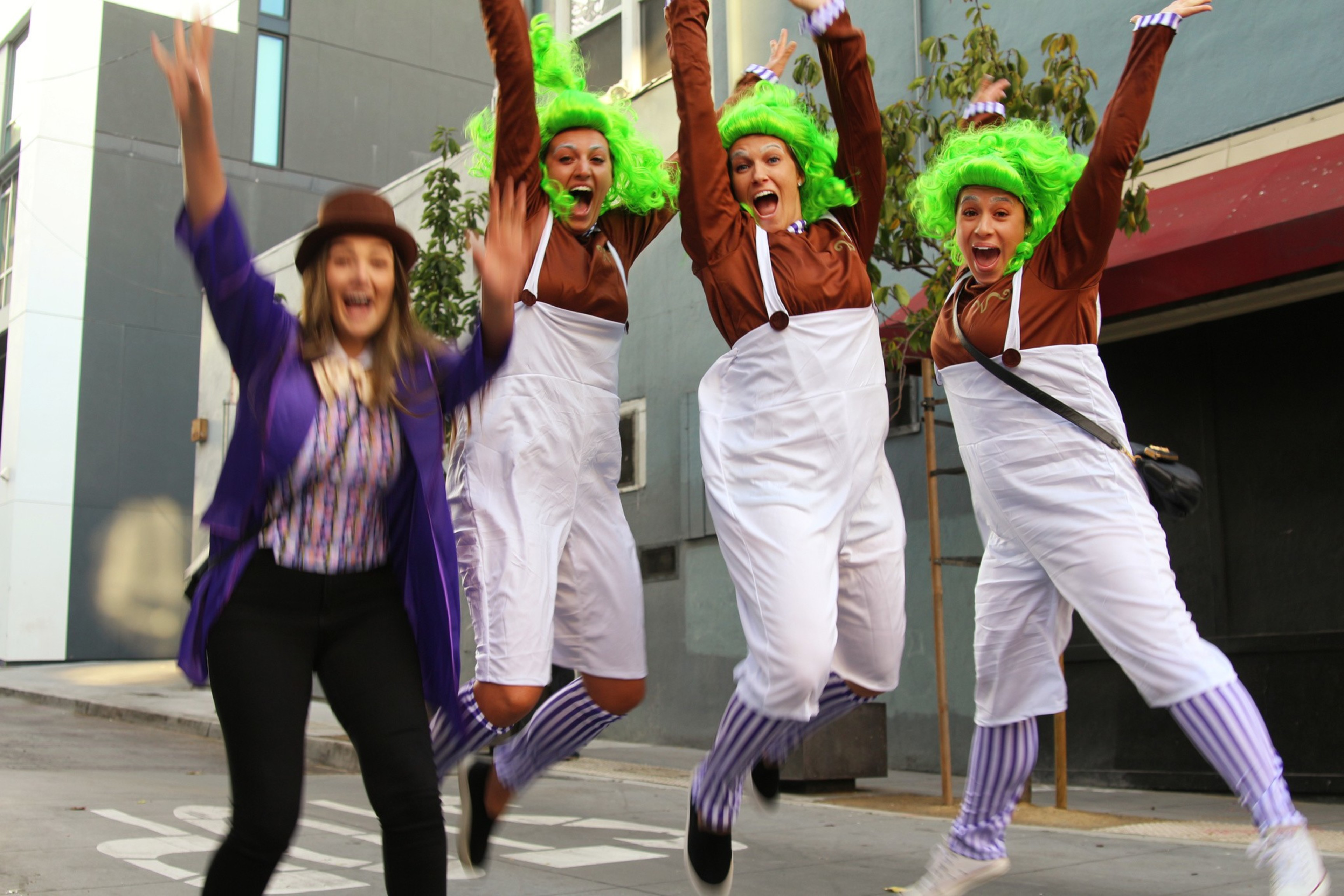 Three people in green wigs and white overalls and one person in a purple jacket and brown hat jump excitedly on a city street.