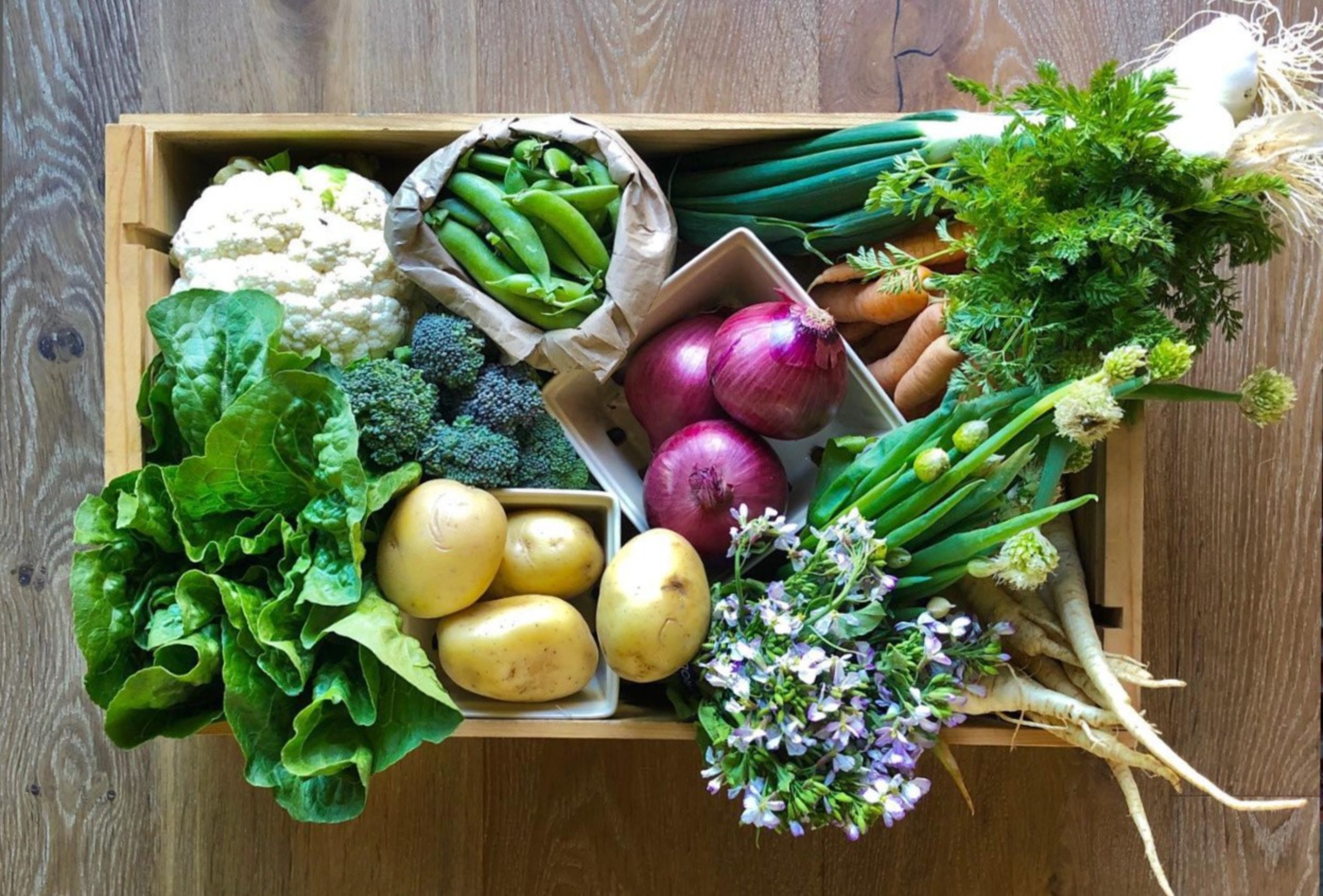 A wooden box filled with fresh vegetables: cauliflower, snap peas, broccoli, leafy greens, potatoes, red onions, herbs, carrots, green onions, and flowers.