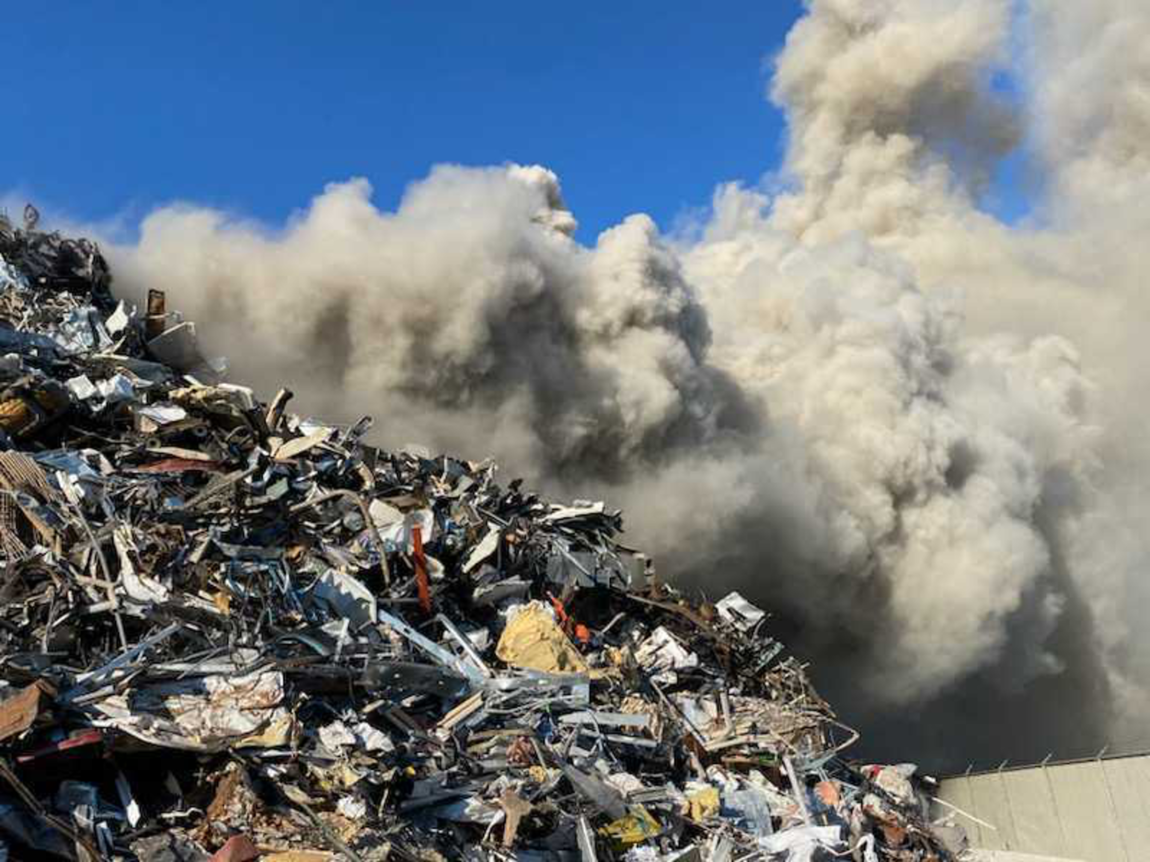 A large pile of metal scrap and debris emits thick, dark smoke against a clear blue sky, suggesting a fire or heavy combustion nearby.
