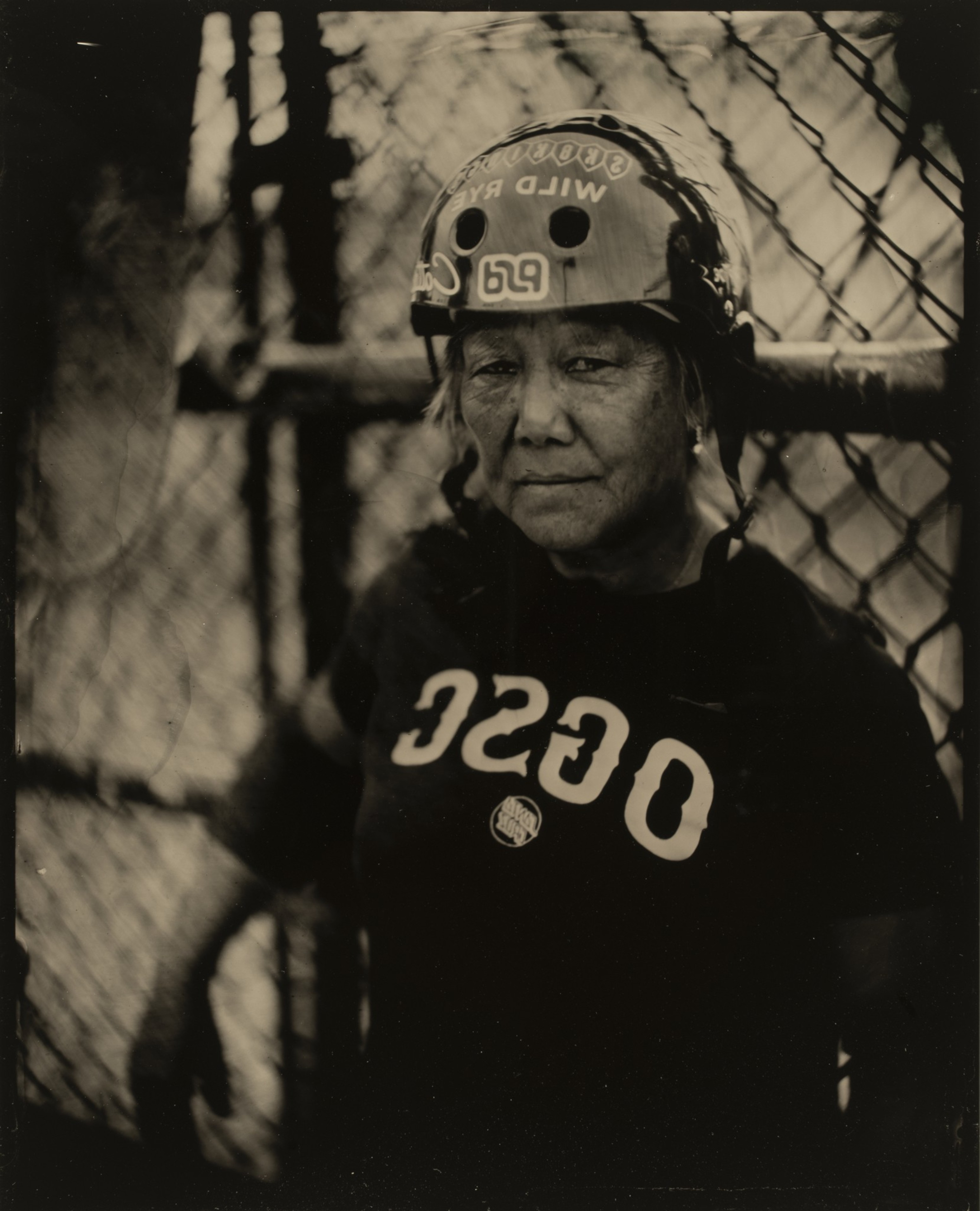 An individual wearing a helmet stands by a chain-link fence, looking directly at the camera with a serious expression. The shirt reads &quot;OSSC.&quot;