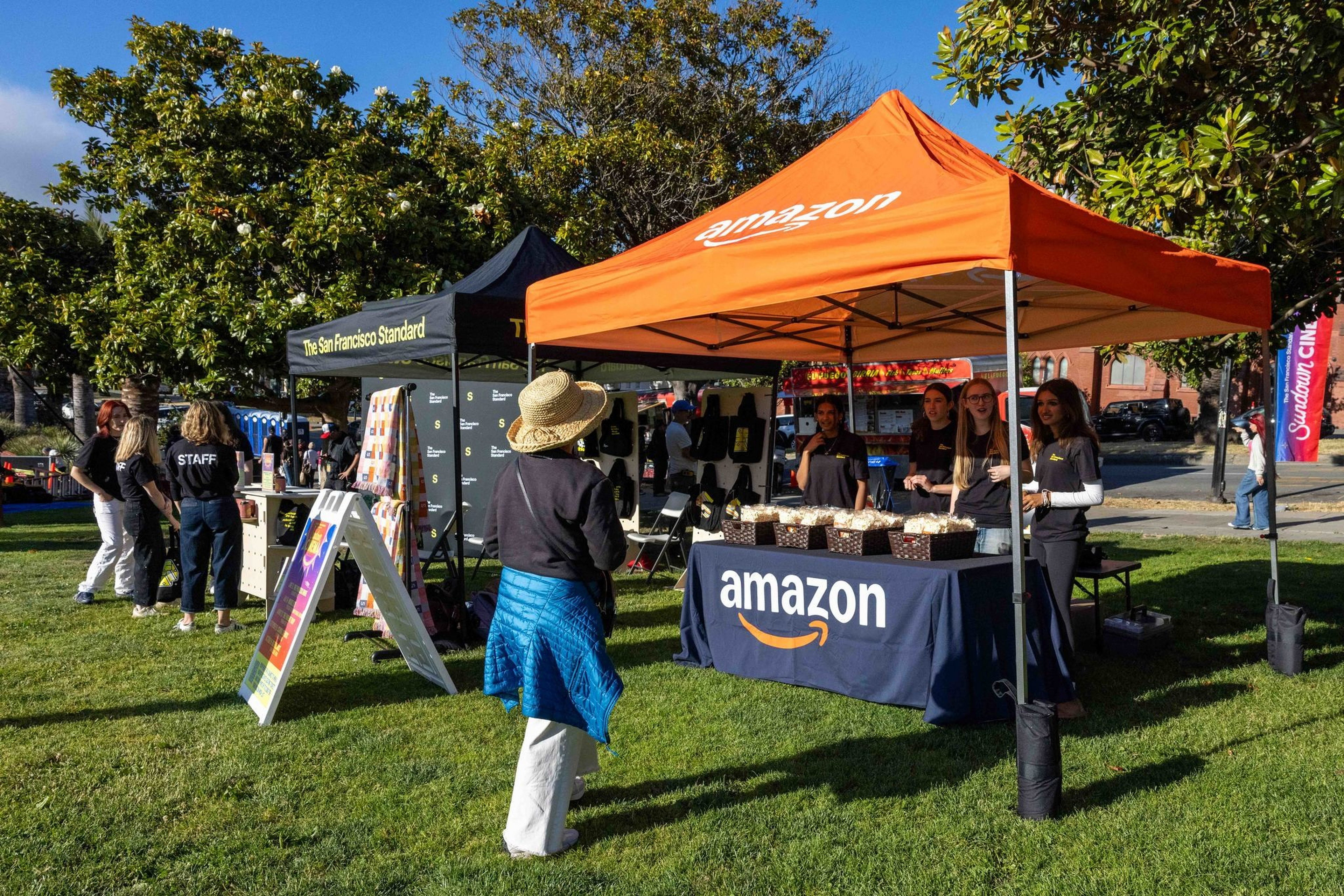 An outdoor event features Amazon and The San Francisco Standard booths, with staff and visitors under sunny skies and green trees on a grassy lawn.
