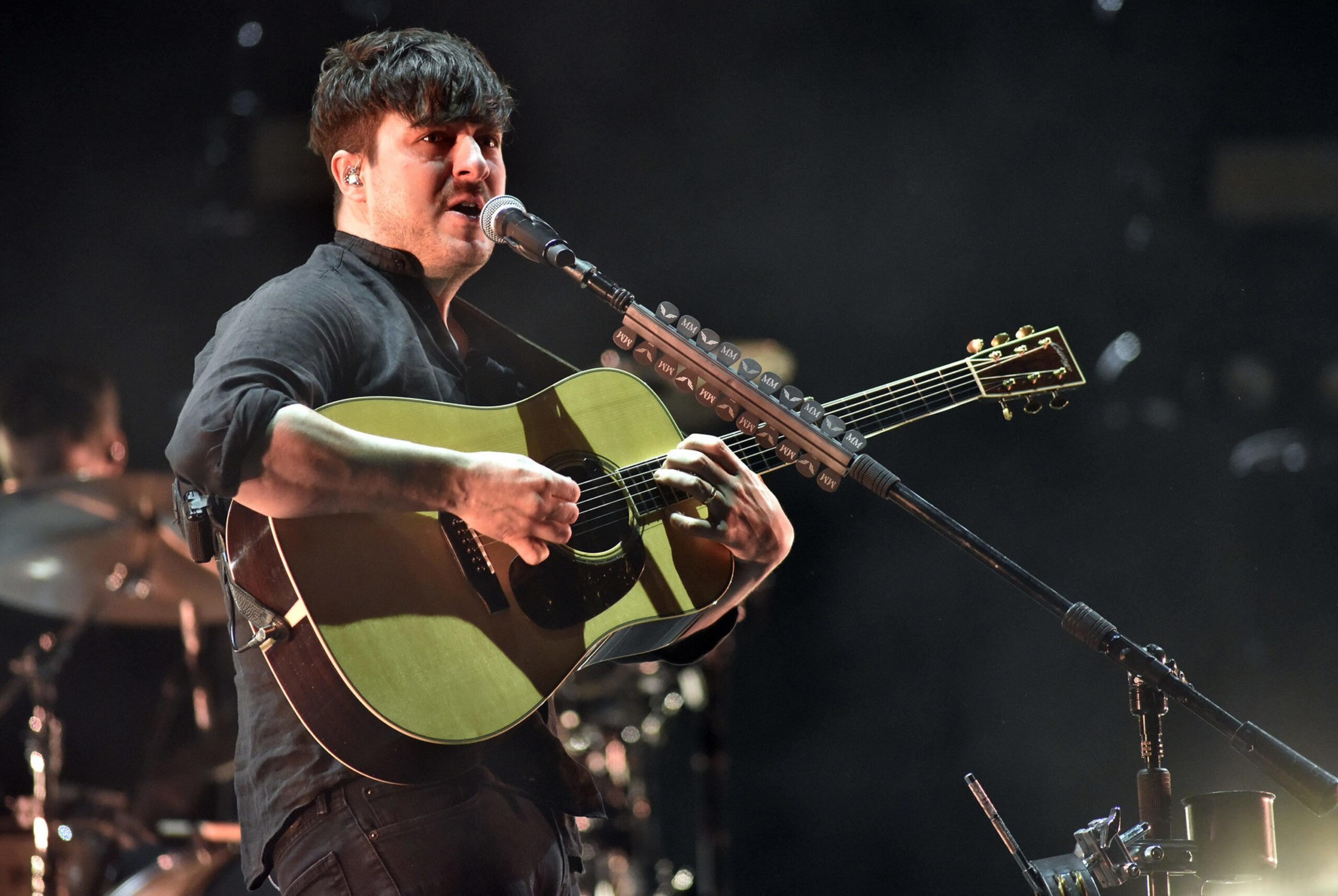 A man wearing a black shirt plays an acoustic guitar and sings into a microphone on a dark stage with drum set in the background.