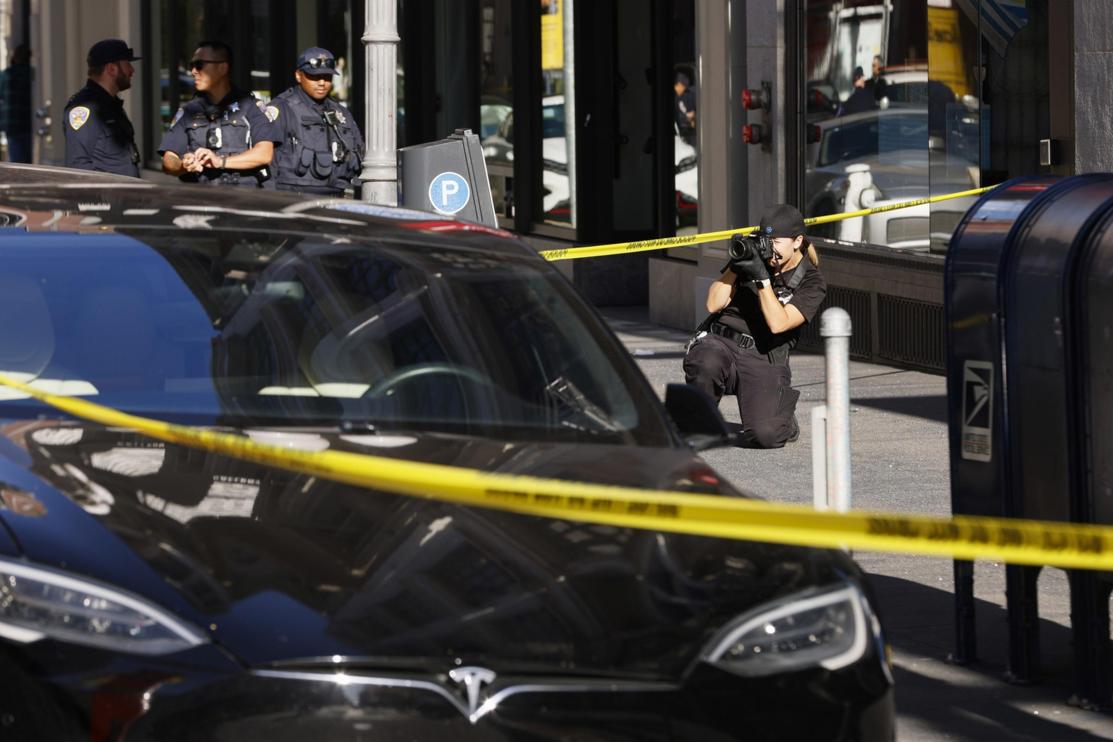 Three police officers are standing near a crime scene, with one officer kneeling and taking photos. Yellow tape cordons off the area around a black Tesla.