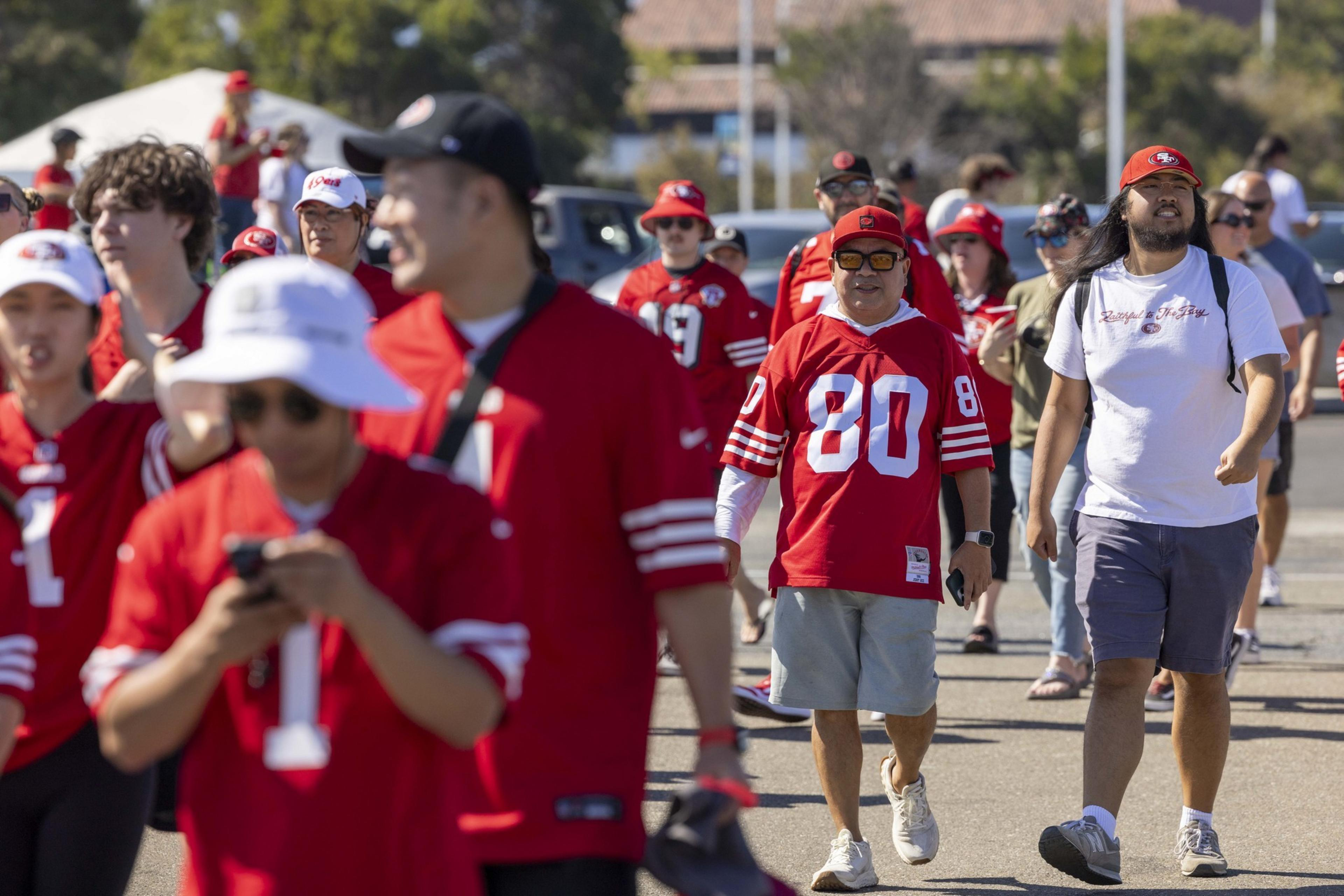 A group of people in red sports jerseys and hats walk outdoors on a sunny day. Some are checking their phones, while others chat and smile.
