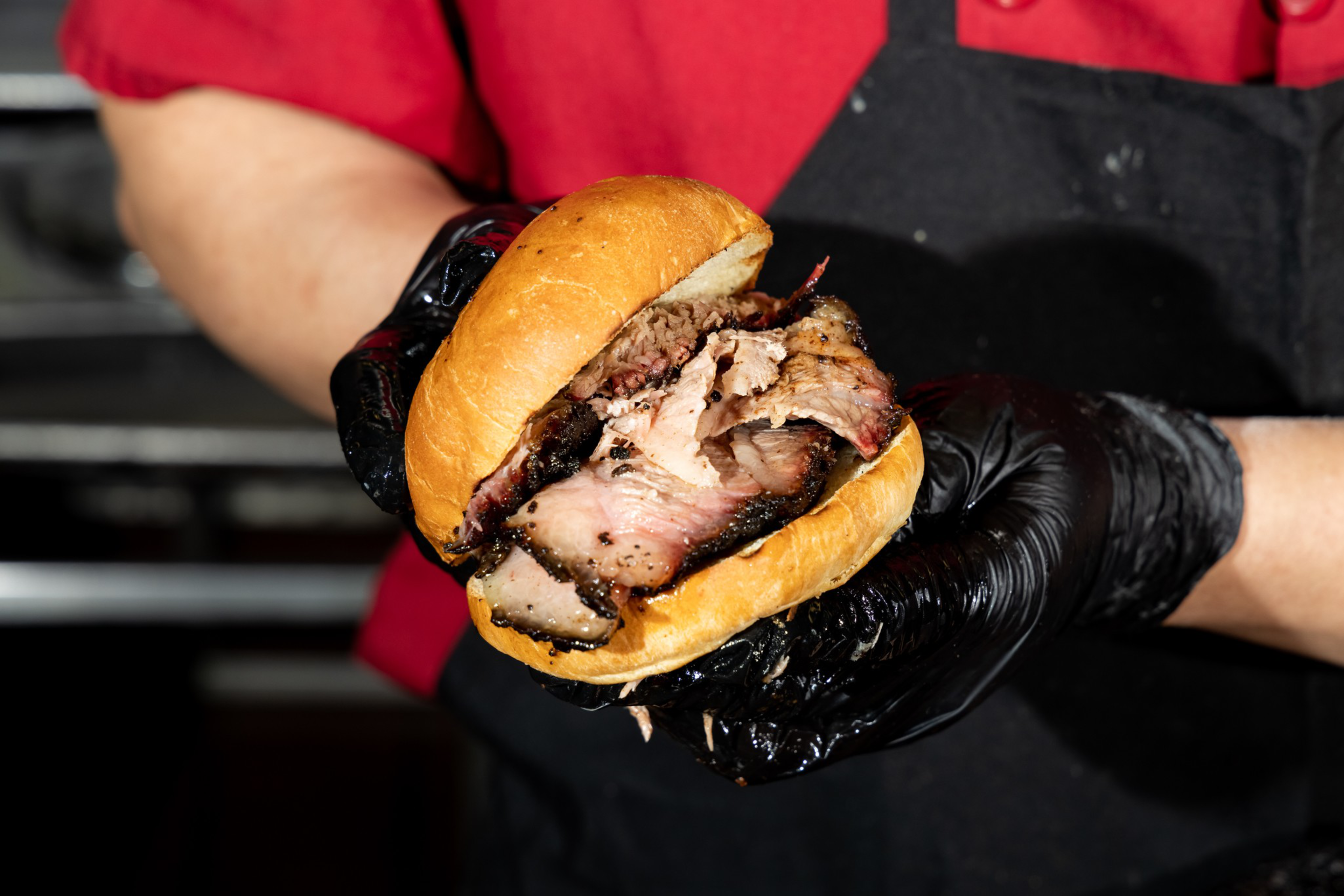 A person wearing black gloves holds a sandwich filled with thick slices of smoked, grilled meat in a soft bun.