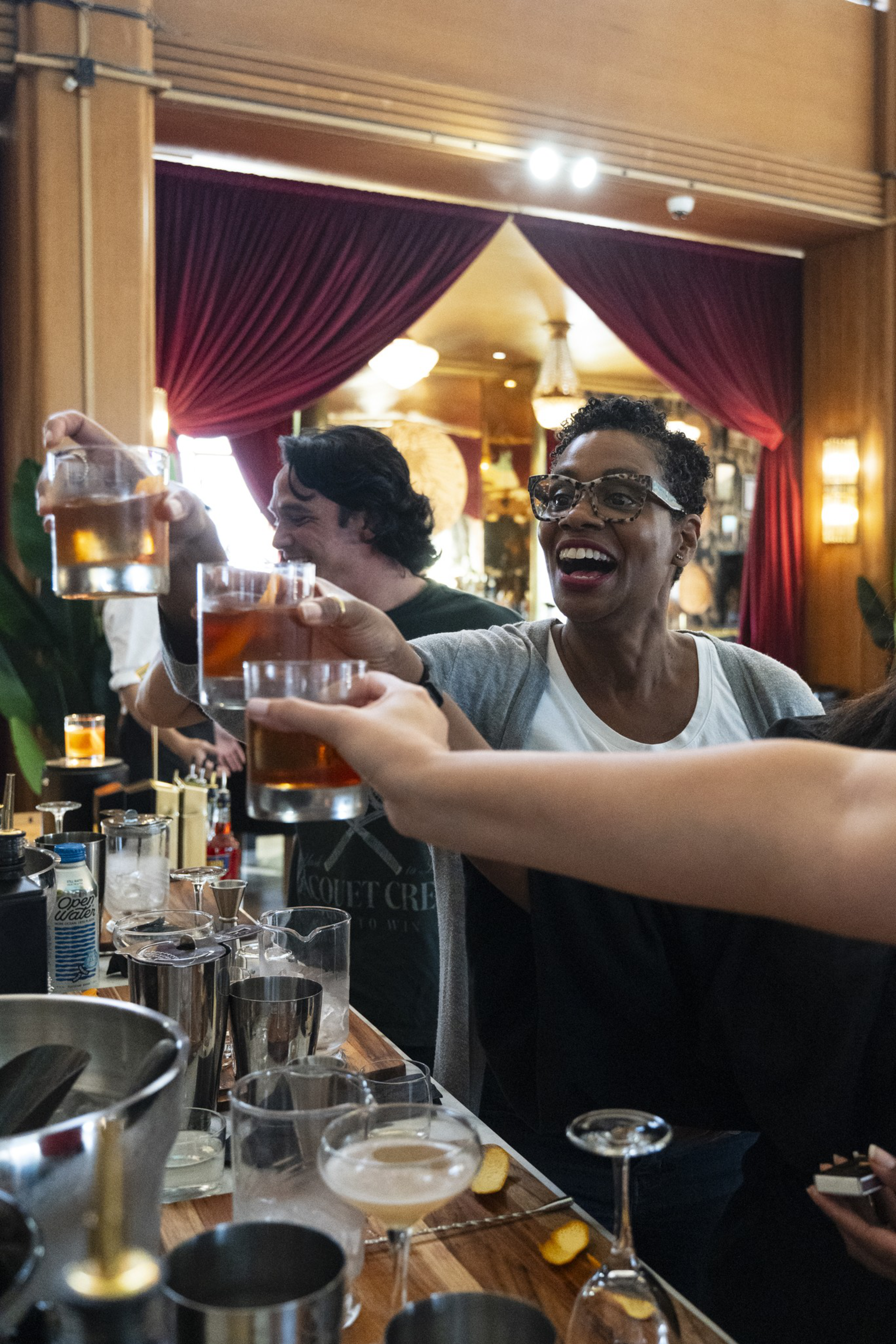A group of people cheerfully clink glasses filled with amber-colored drinks at a bar with red curtains and warm lighting.
