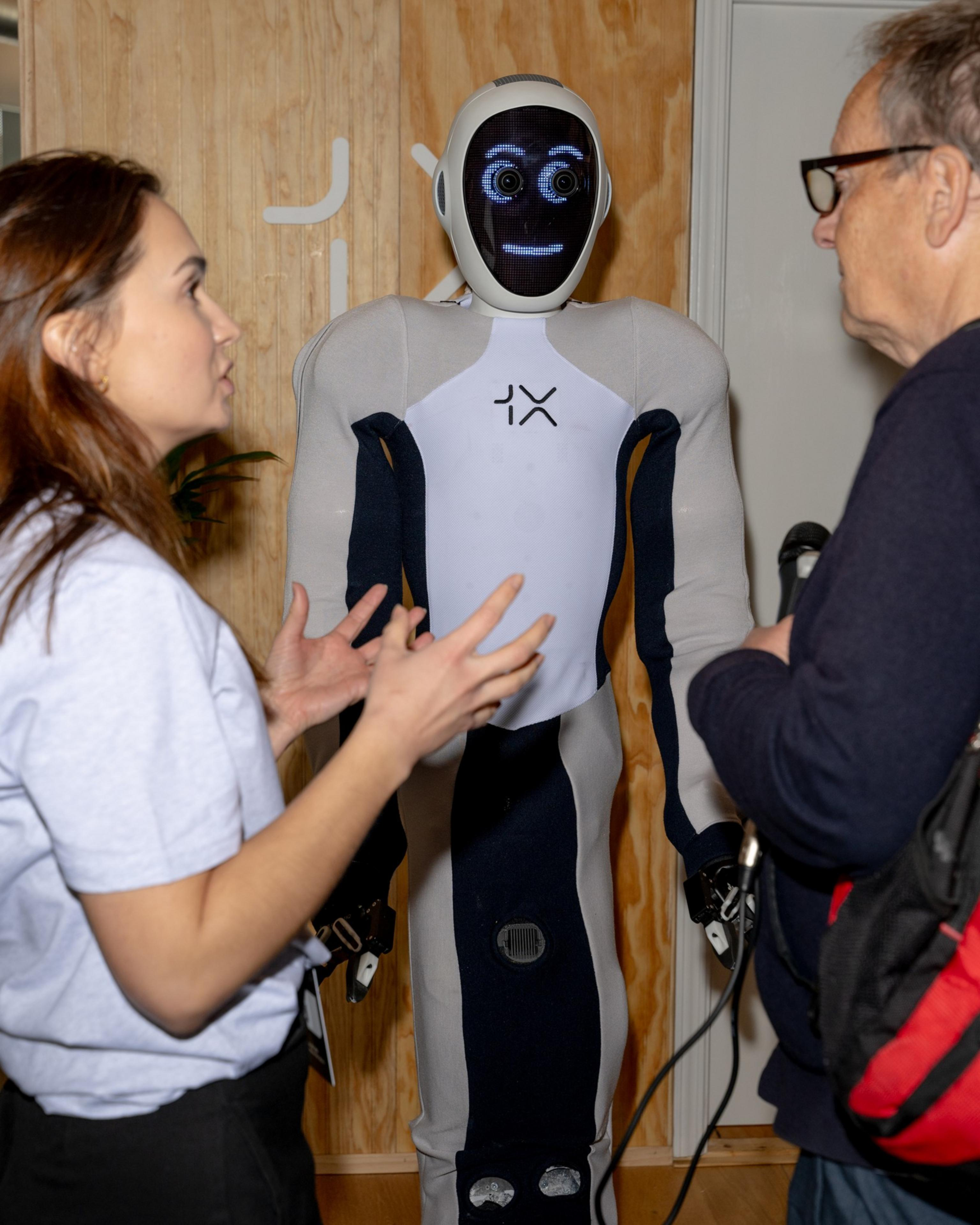 A humanoid robot with expressive eyes stands between a woman talking animatedly and a man holding a microphone. The setting is indoors, with a wooden wall.