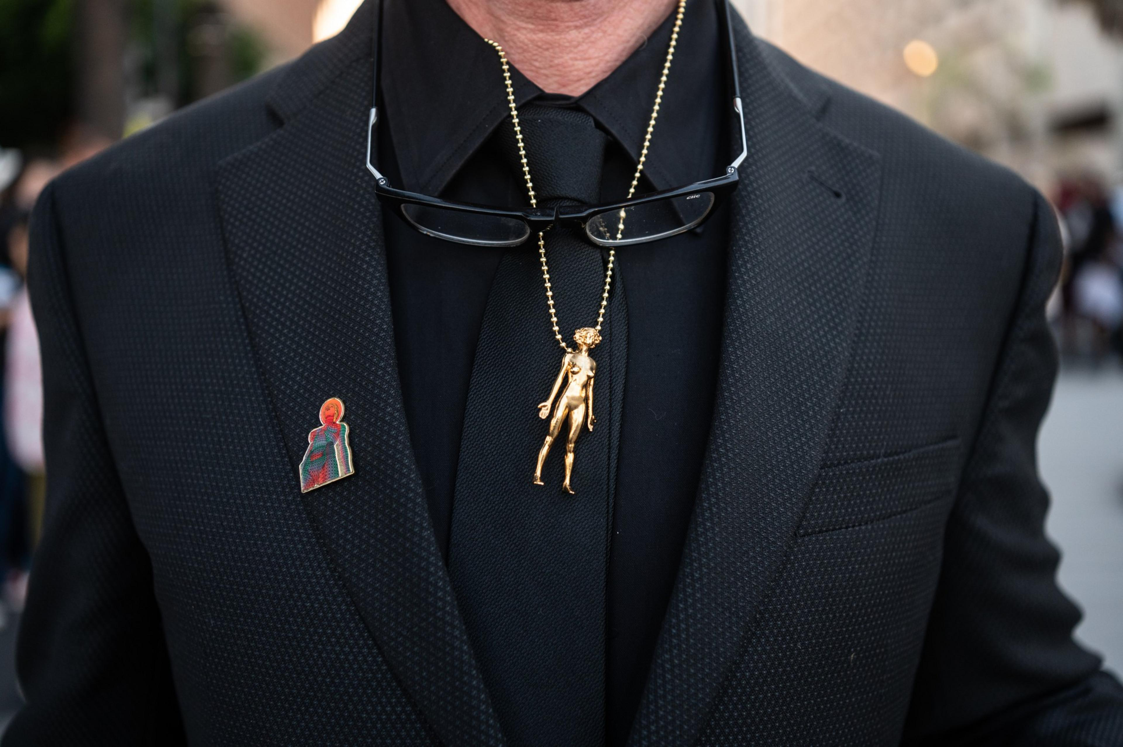 A man in a black suit wears glasses on his neck and a golden humanoid pendant. A colorful pin of a figure in a helmet is on the suit's lapel.