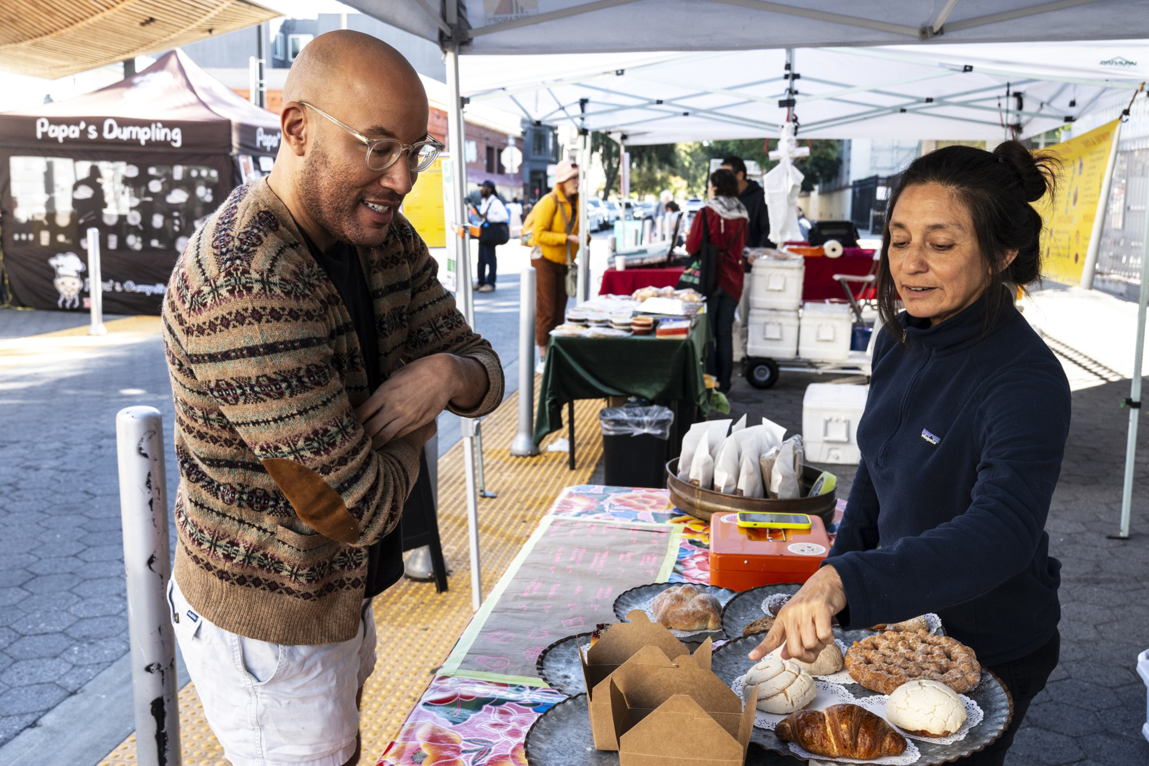 A woman behind a market stall points to baked goods while a man in glasses and a patterned sweater looks on with interest.