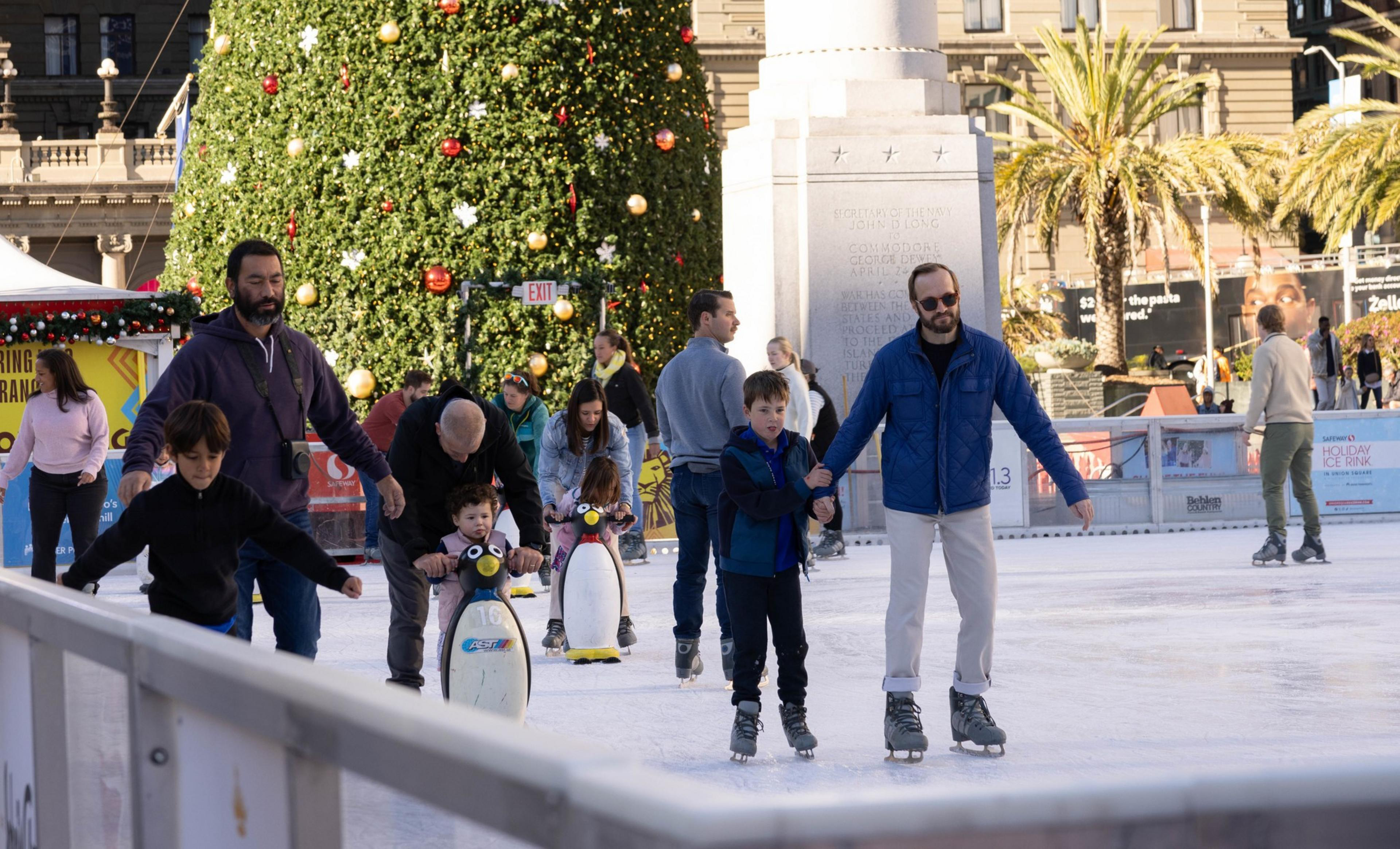 People skate on an ice skating rink with a Christmas tree behind them decorated with red and white bulbs.