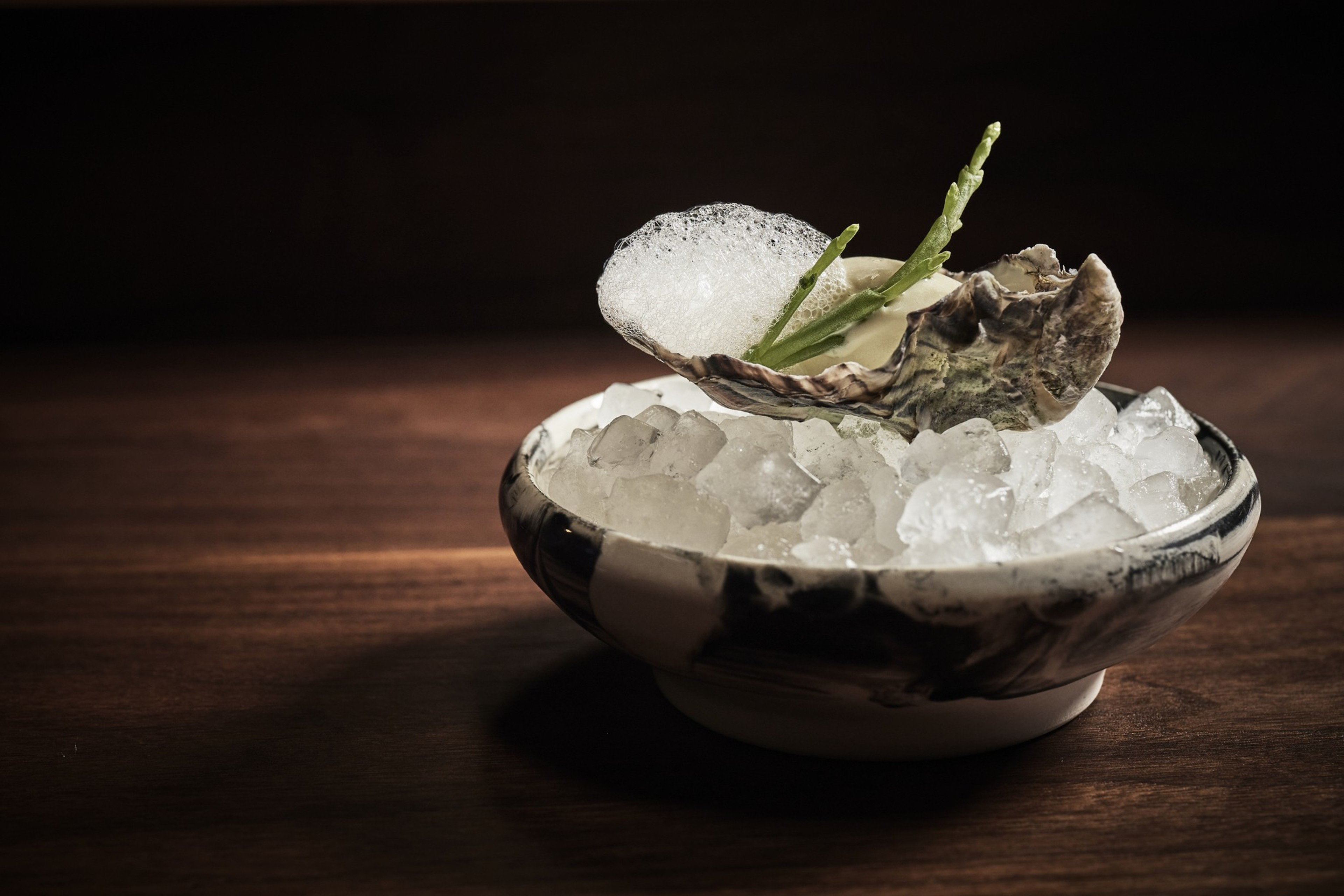 A cracked oyster shell with foam and green garnish rests on crushed ice in a black and white bowl on a wooden surface.