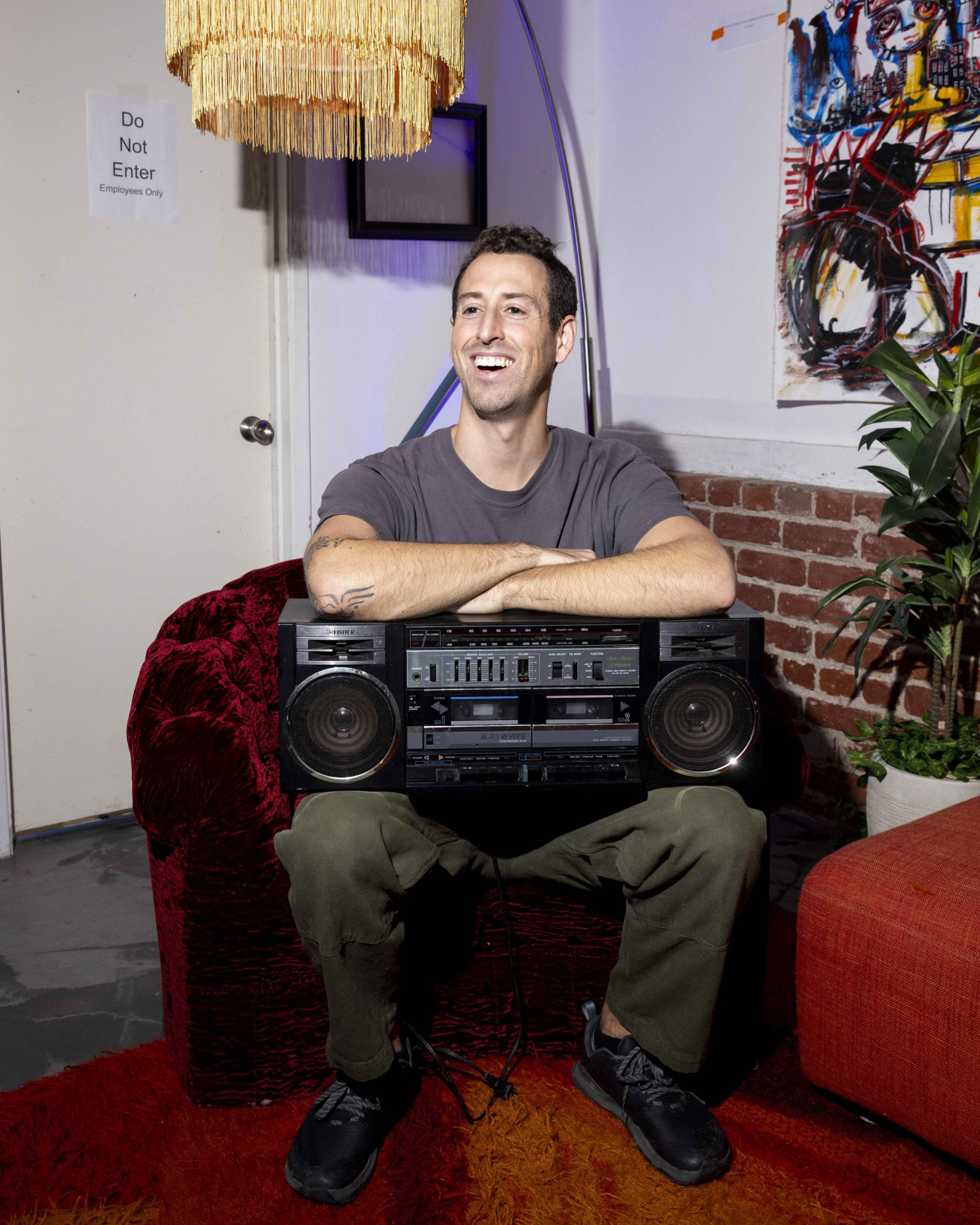 A man smiles while sitting on a red chair, holding a large boombox. There's a fringed lamp, a &quot;Do Not Enter&quot; sign, abstract wall art, and a potted plant nearby.
