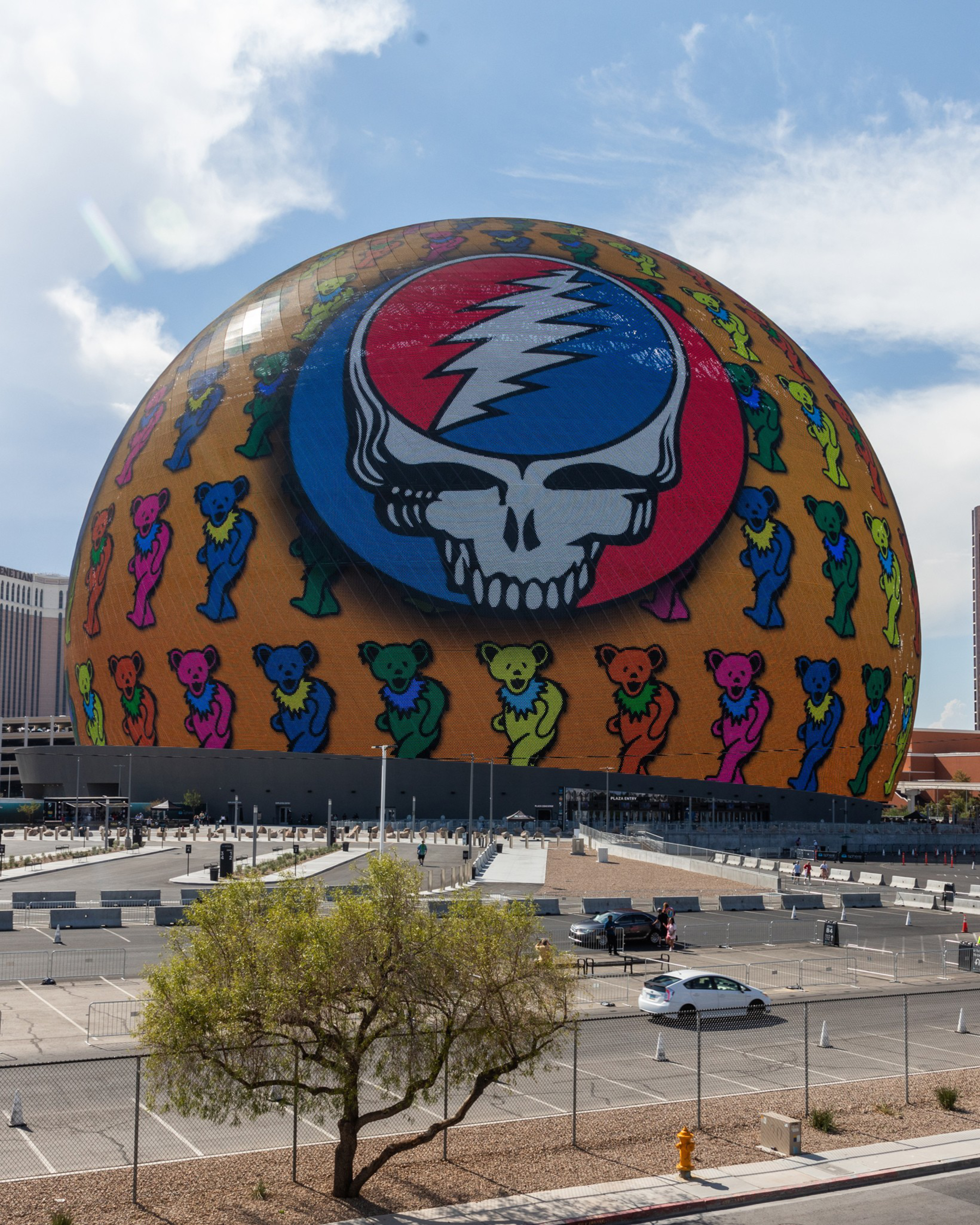 A large, round building displays a colorful graphic featuring a skull with a lightning bolt and dancing bears. The foreground includes a parking lot and a tree.