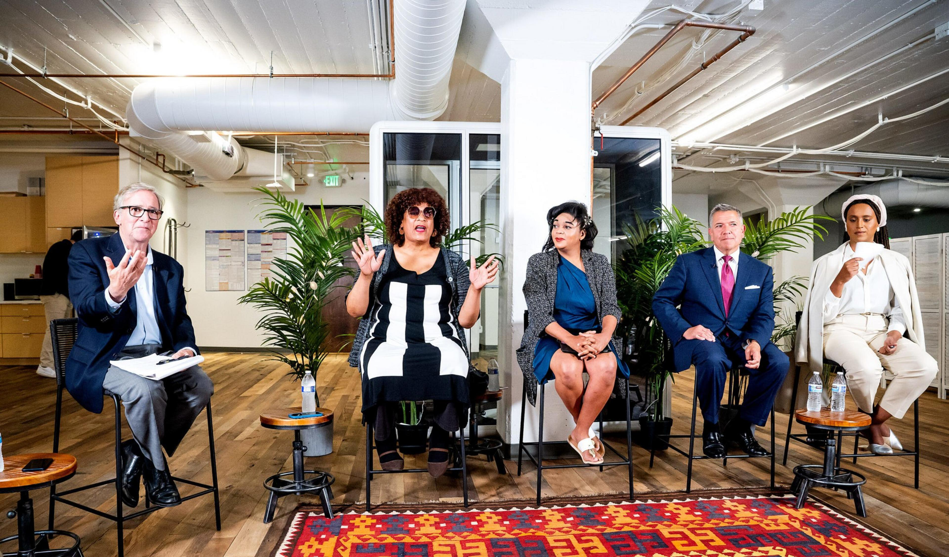 Five people, three women and two men, sit on high stools in a modern office space, engaged in a discussion with plants and a colorful rug nearby.