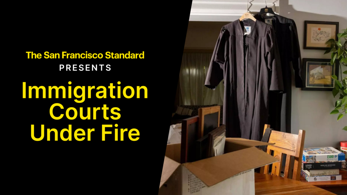 Two black judicial robes hang on hangers in a dimly lit room with a wooden dining table, chairs, a cardboard box, and framed pictures on the wall.