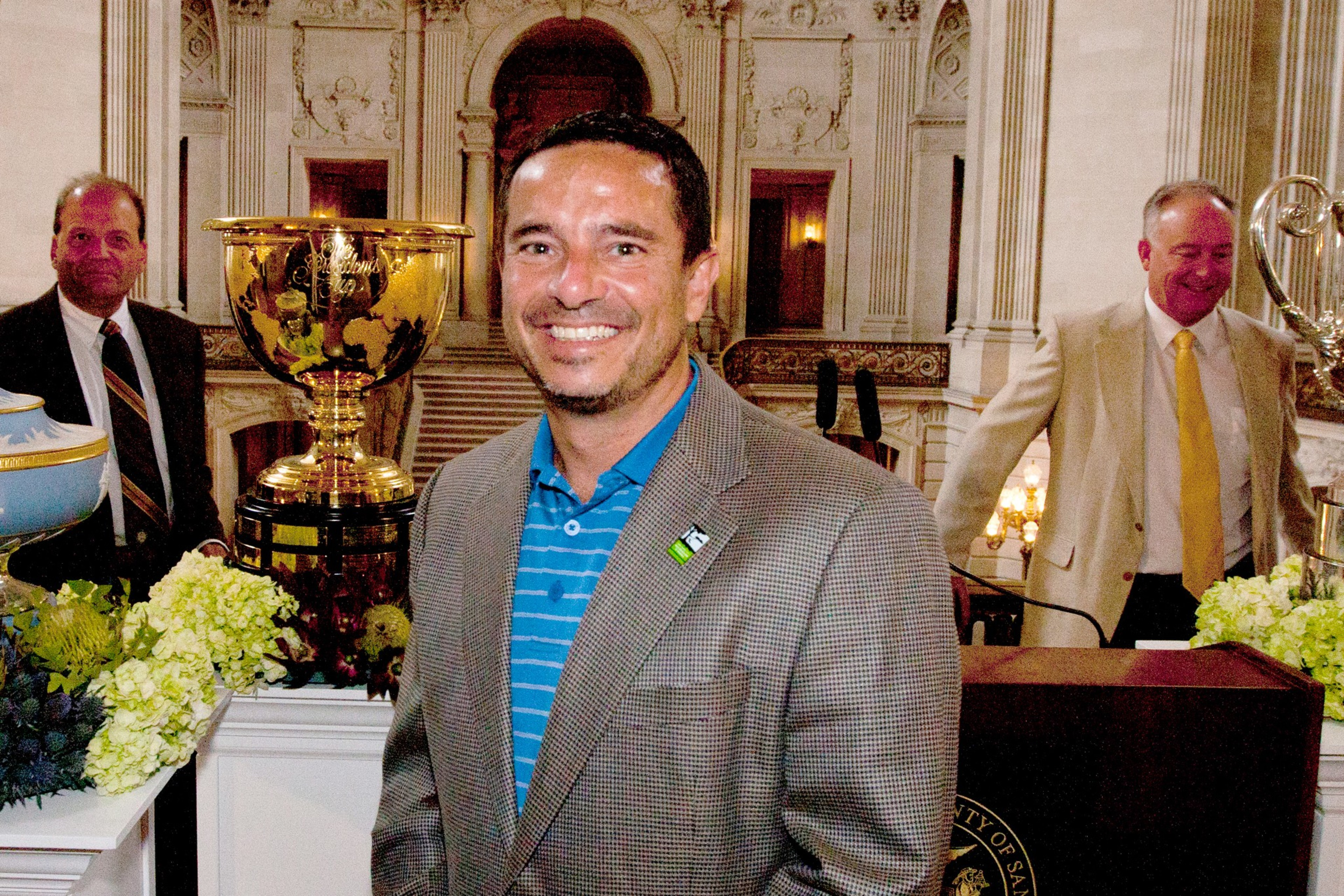 A man in a checkered jacket and blue striped shirt smiles in front of large trophies, with two men standing behind him near floral arrangements.