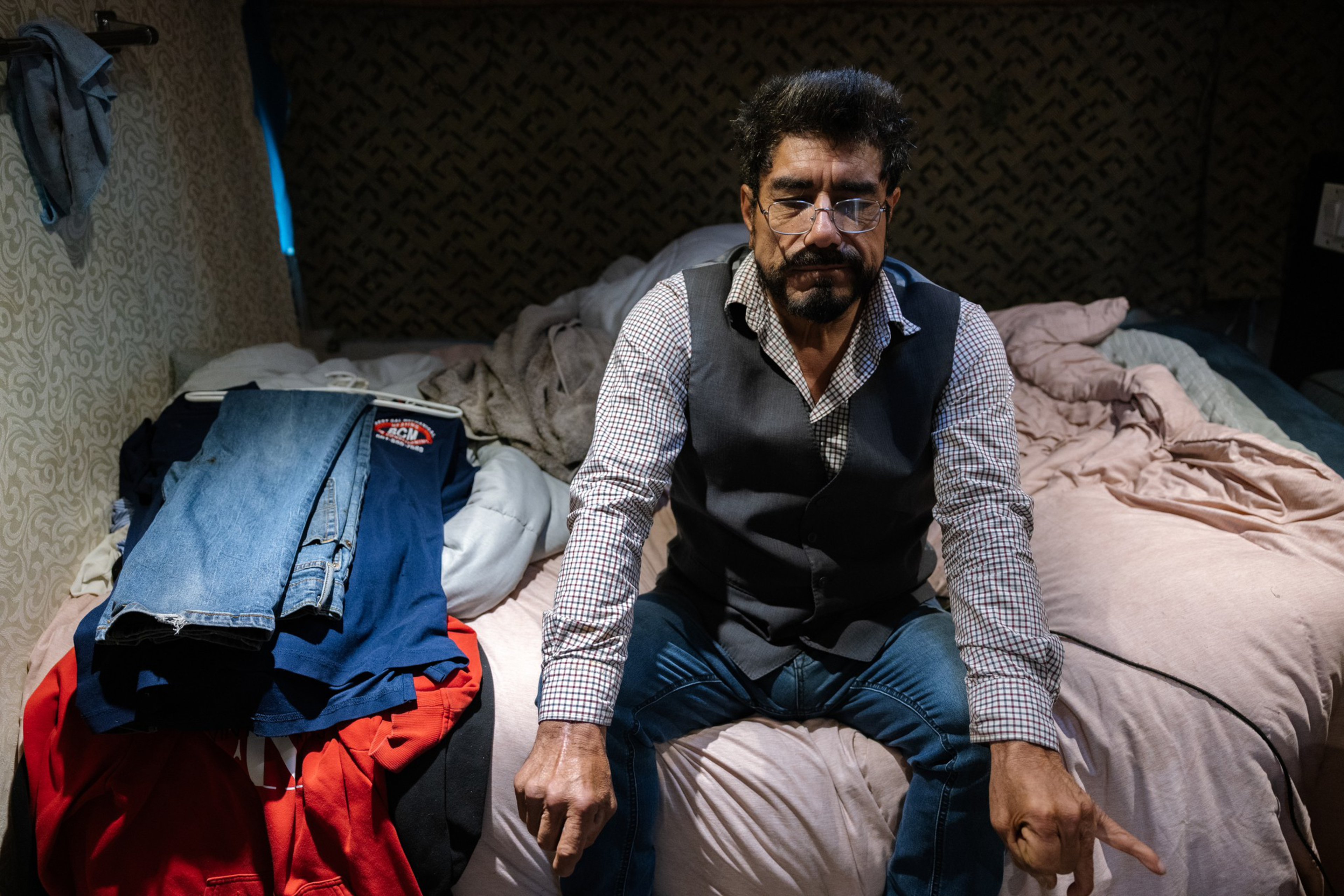 A man with glasses and a beard sits on a bed with messy blankets, wearing a checkered shirt and vest, pointing down with both hands.
