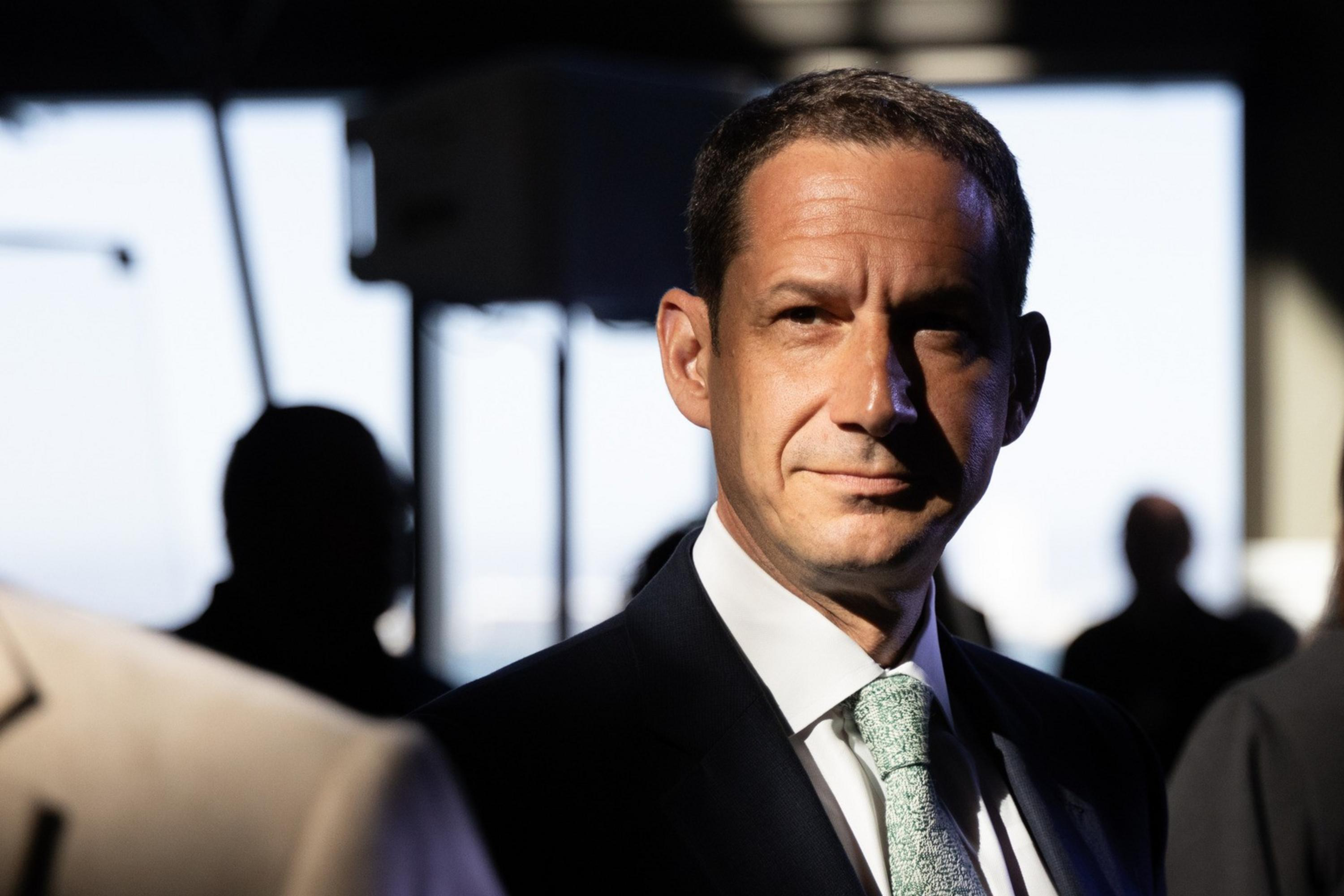 A man in a suit and tie stands in dramatic light with shadows on his face, surrounded by people in a dimly lit indoor setting.