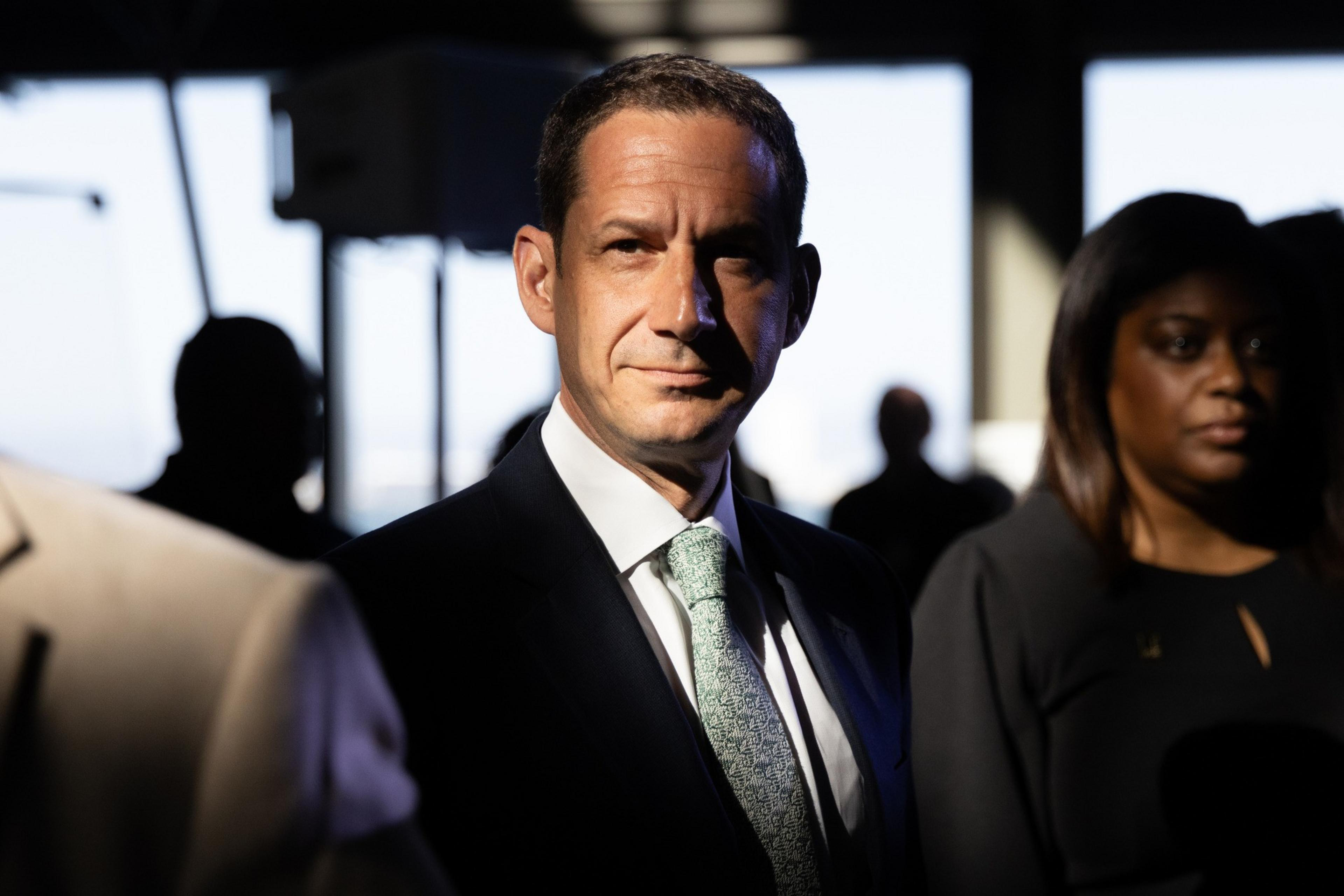A man in a suit and tie stands in dramatic light with shadows on his face, surrounded by people in a dimly lit indoor setting.