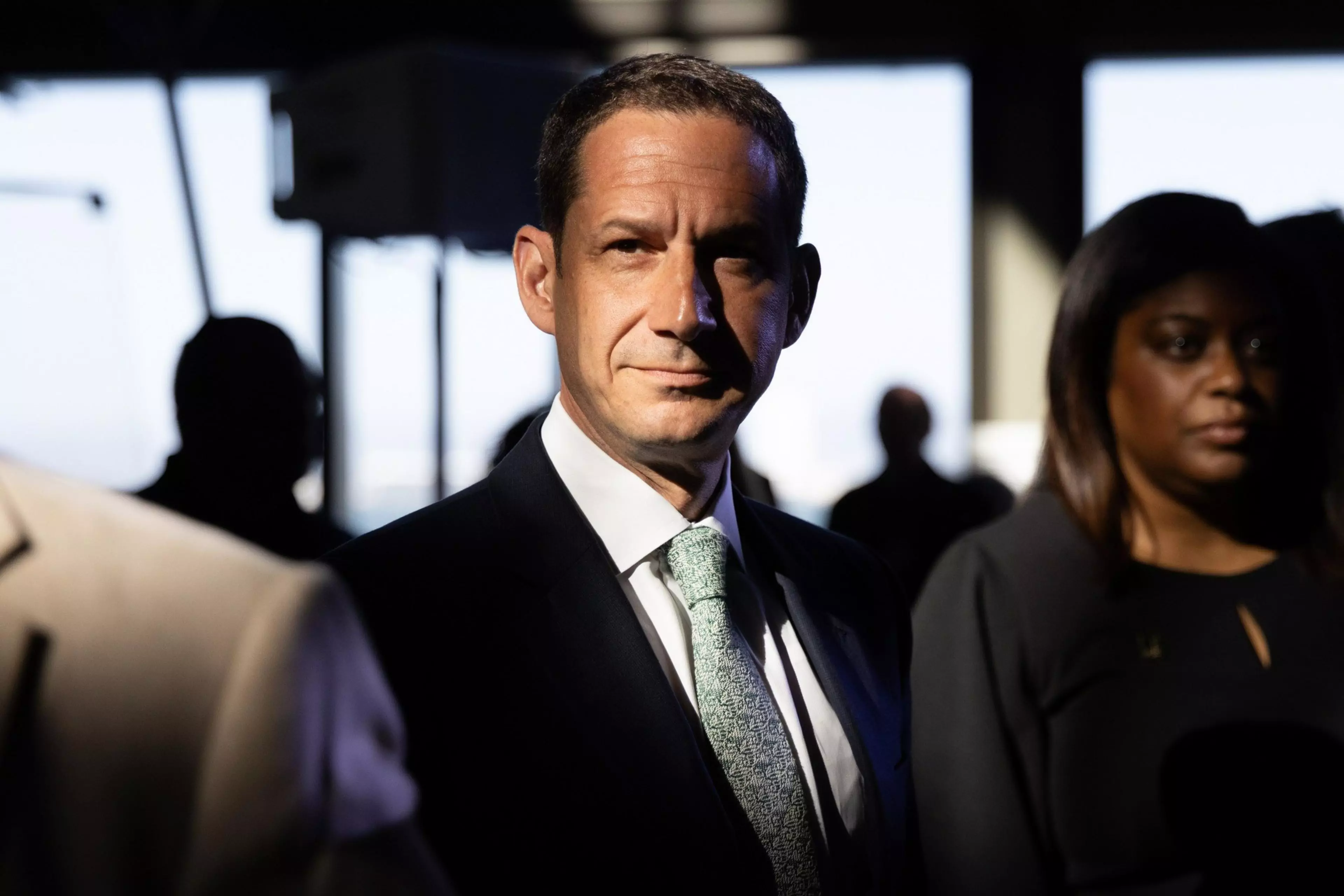 A man in a suit and tie stands in dramatic light with shadows on his face, surrounded by people in a dimly lit indoor setting.