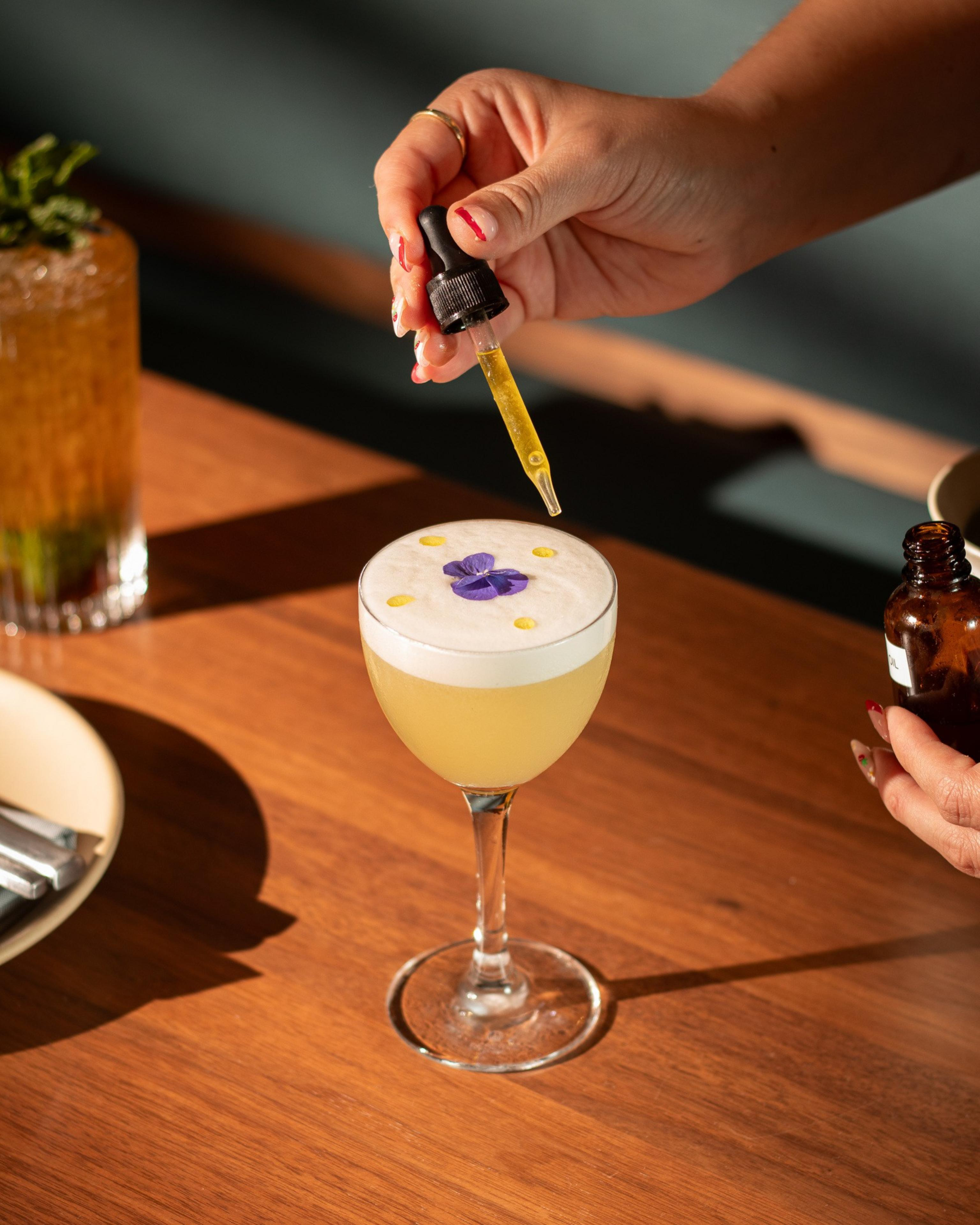 A hand holds a dropper adding yellow droplets onto a frothy cocktail garnished with a purple edible flower, placed on a wooden table.
