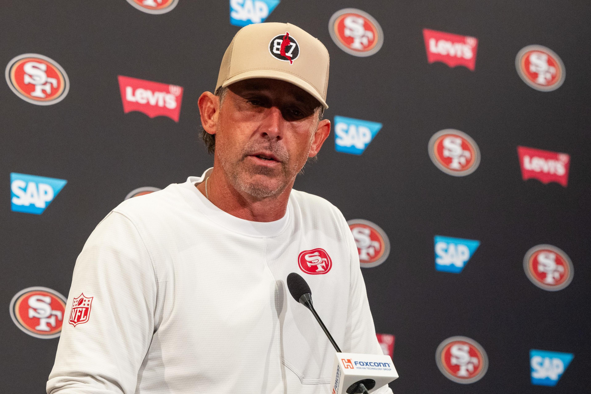 A man wearing a white San Francisco 49ers sweatshirt and a tan cap speaks at a press conference with a microphone in front of him.