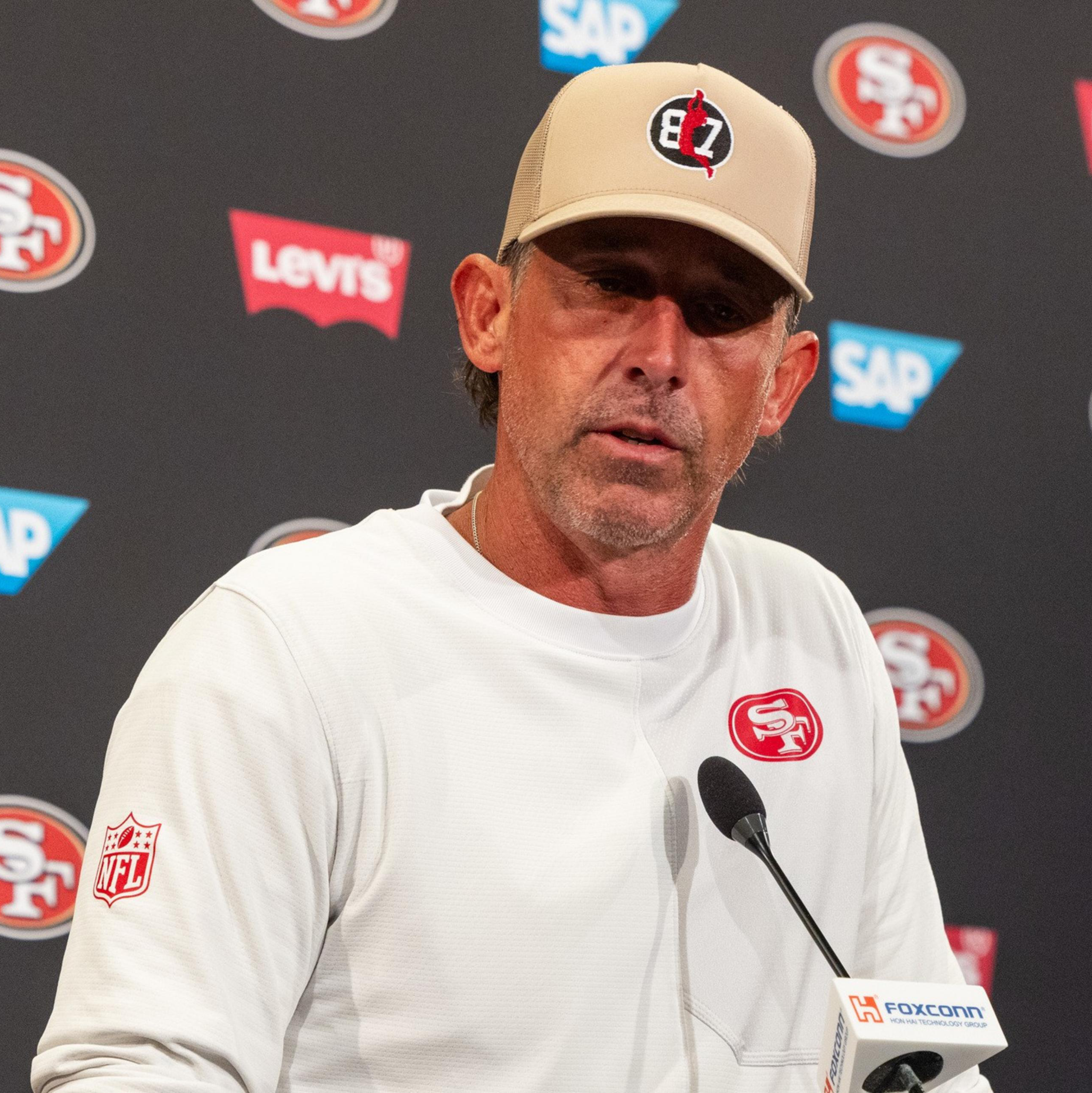 A man wearing a white San Francisco 49ers sweatshirt and a tan cap speaks at a press conference with a microphone in front of him.