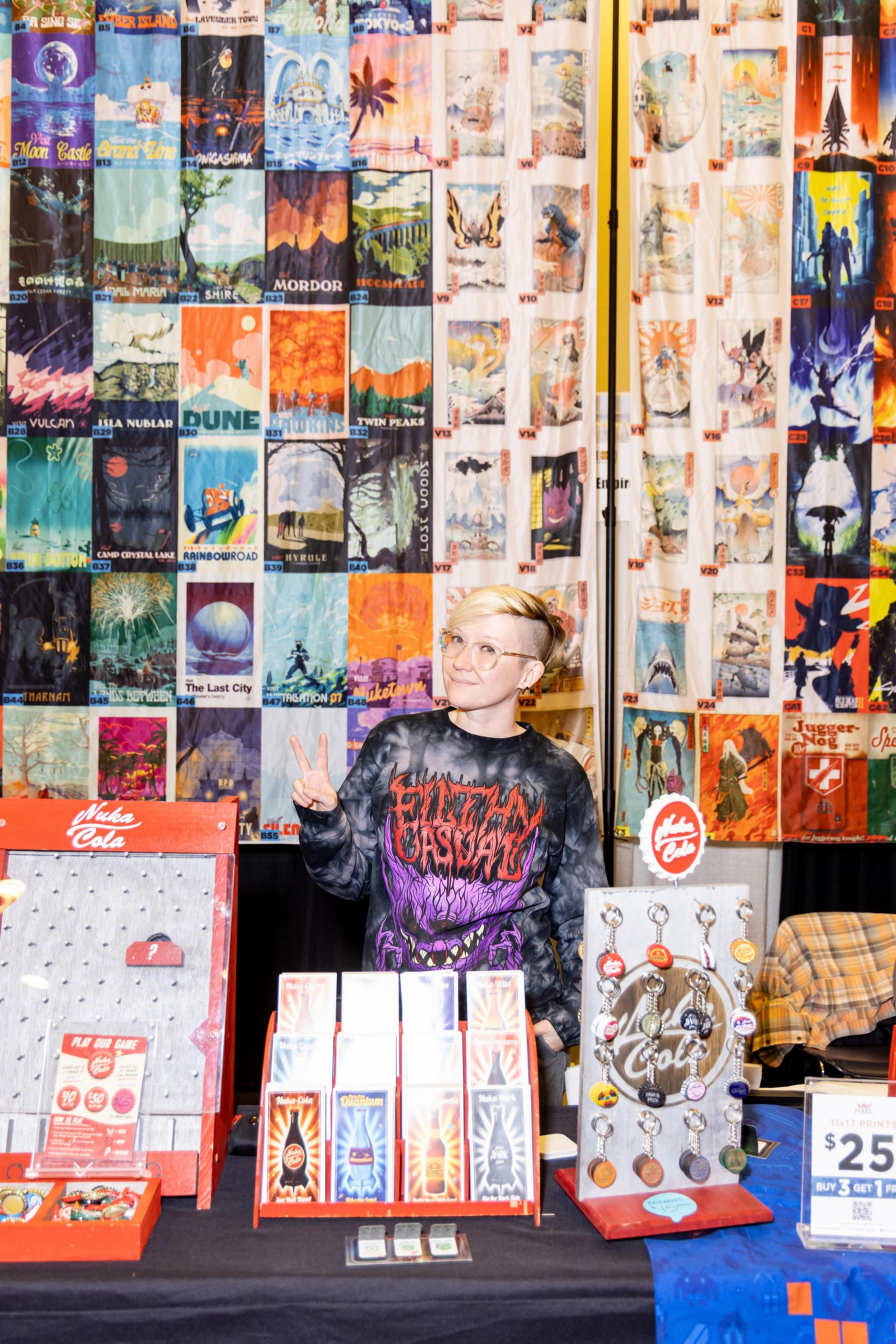A person stands at a booth with colorful posters and merchandise, giving a peace sign. The setup includes keychains, cards, and a Nuka Cola theme.