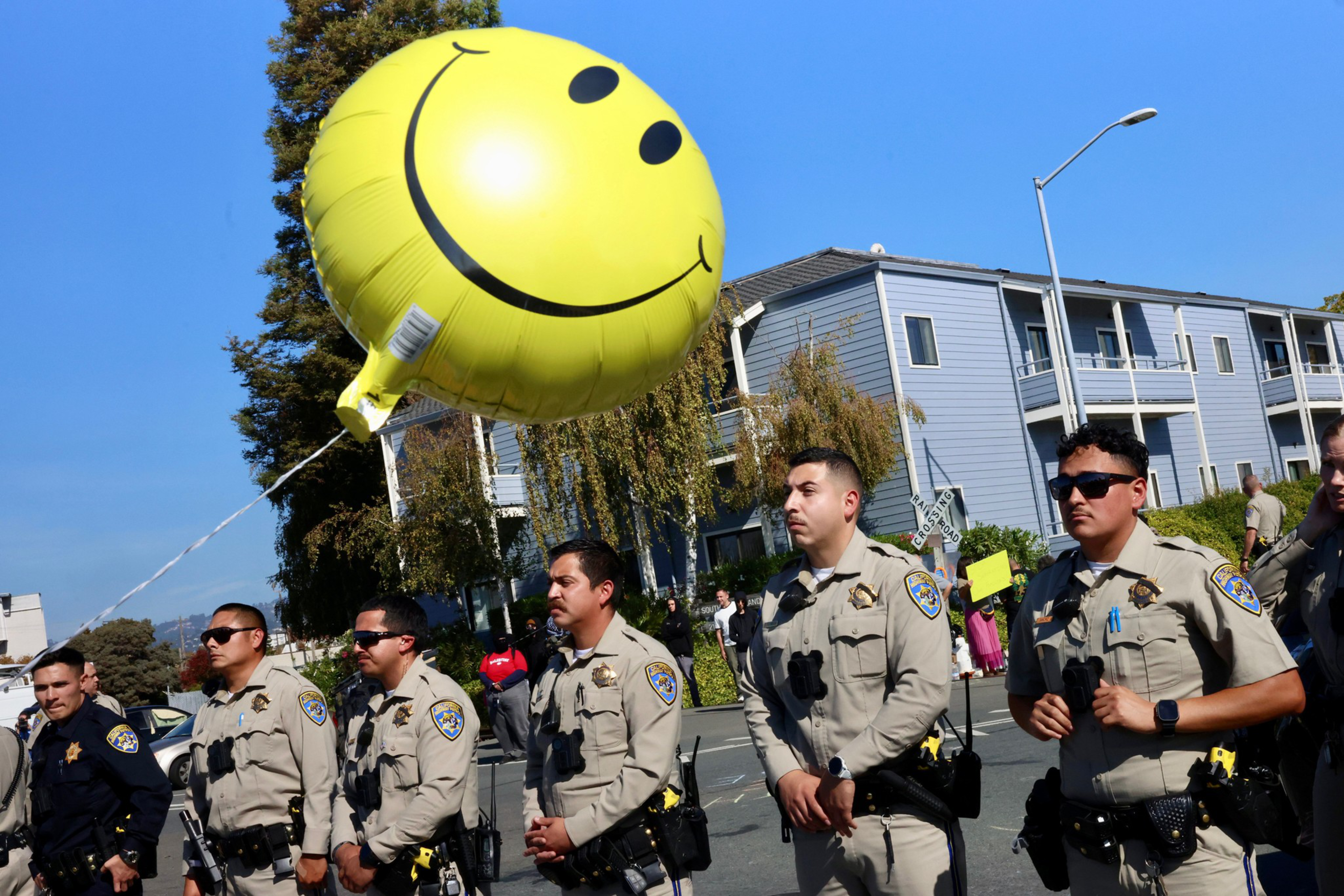 Six police officers stand in a line outdoors near a large yellow smiley face balloon floating above them.