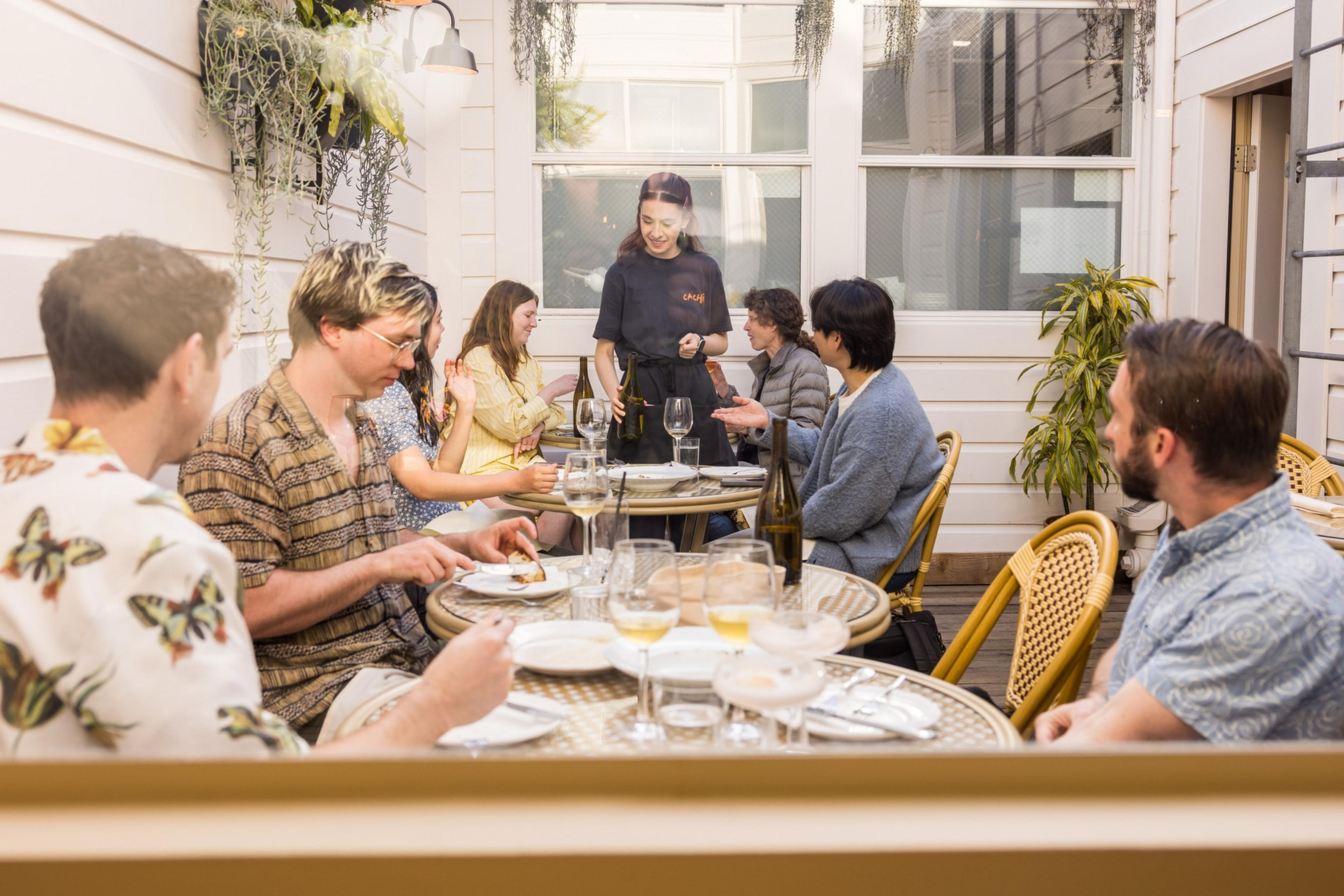 A group of people dine outdoors at a restaurant. They're seated at a round table with drinks and plates. A server in a black outfit is attending to them.